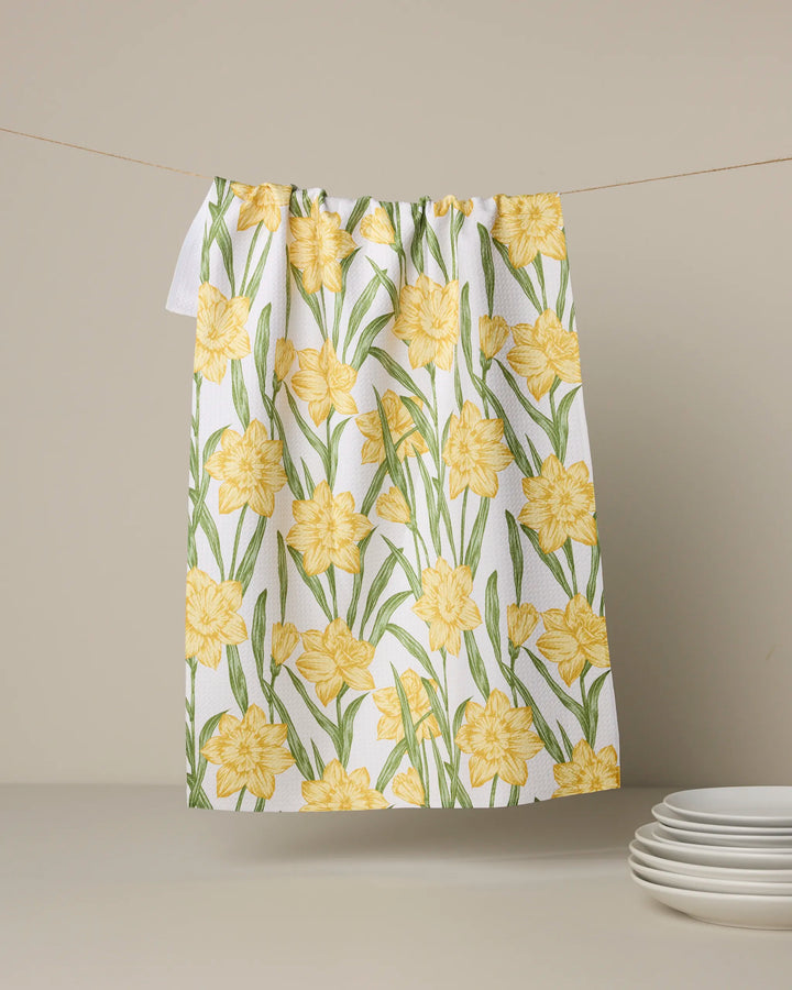 The Geometry "Daffodil Darling" recycled kitchen towel, featuring yellow daffodils and green leaves, hangs on a line against a beige background with a stack of white plates placed below.