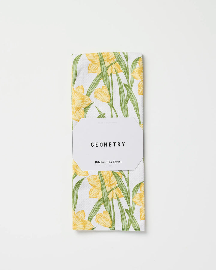 A folded, quick-drying tea towel with a yellow floral and green stem design, called Daffodil Darling by Geometry, is wrapped in a white label reading "GEOMETRY Kitchen Tea Towel" and set against a plain white background.
