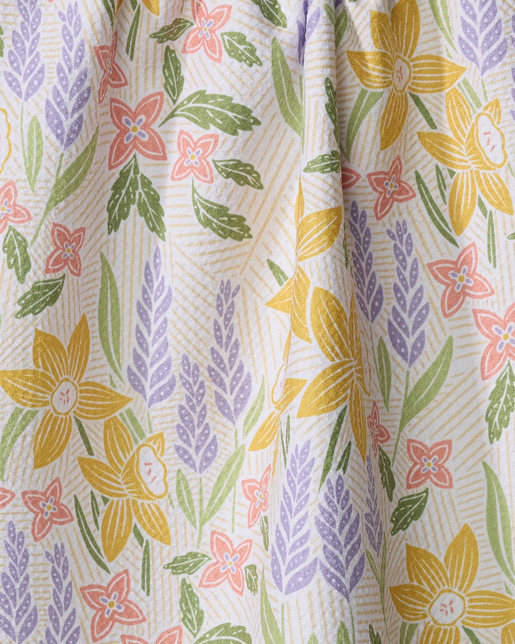 A close-up of the "Daffodil Dreams" tea towel by Sarah Daugherty—vibrant yellow, pink, and purple florals with green leaves and diagonal lines on a light background. Made from quick-drying recycled materials.