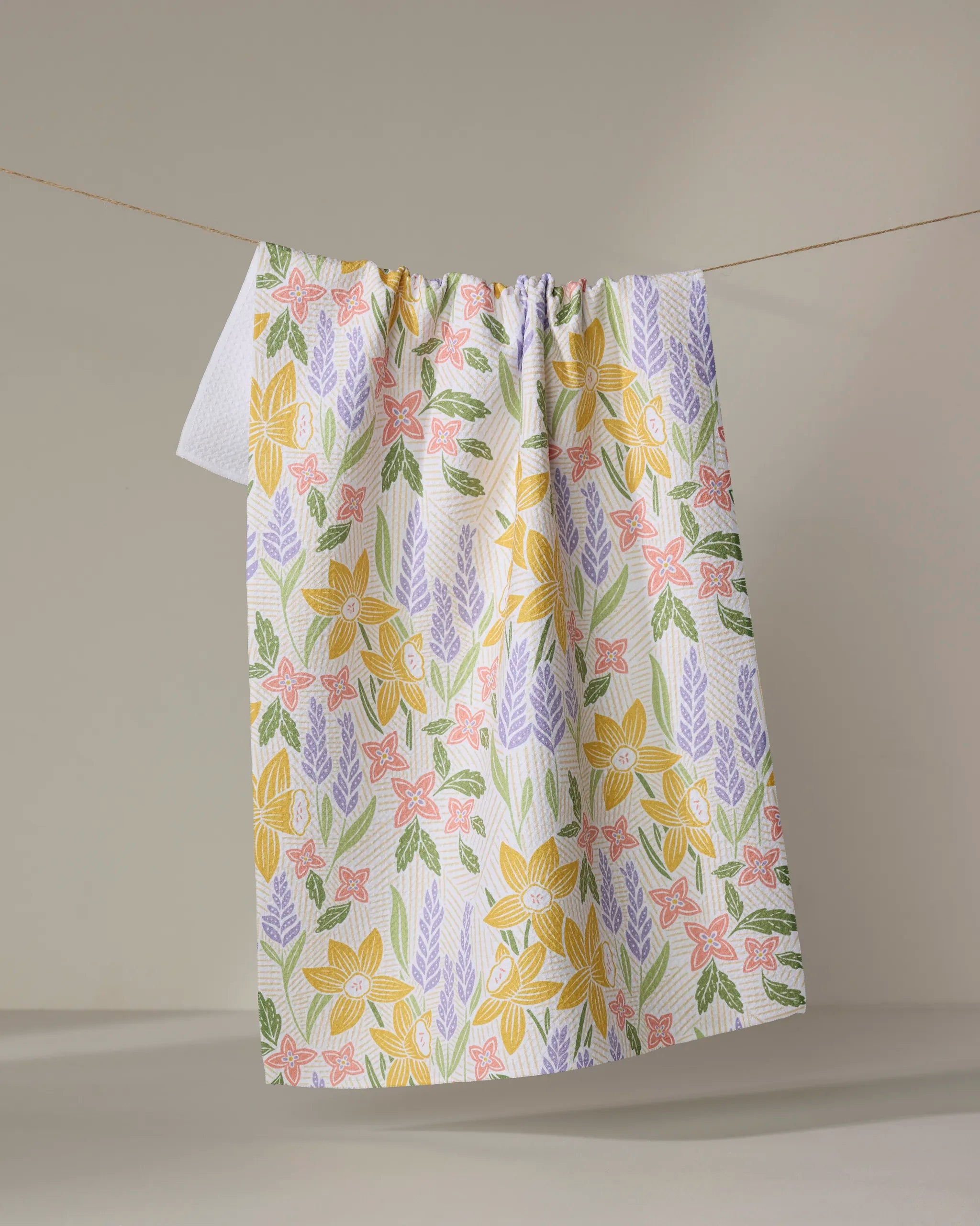 The "Daffodil Dreams" kitchen towel by Sarah Daugherty, made from white recycled fabric with yellow, pink, and purple floral patterns and green leaves, hangs draped over a line against a plain light background.