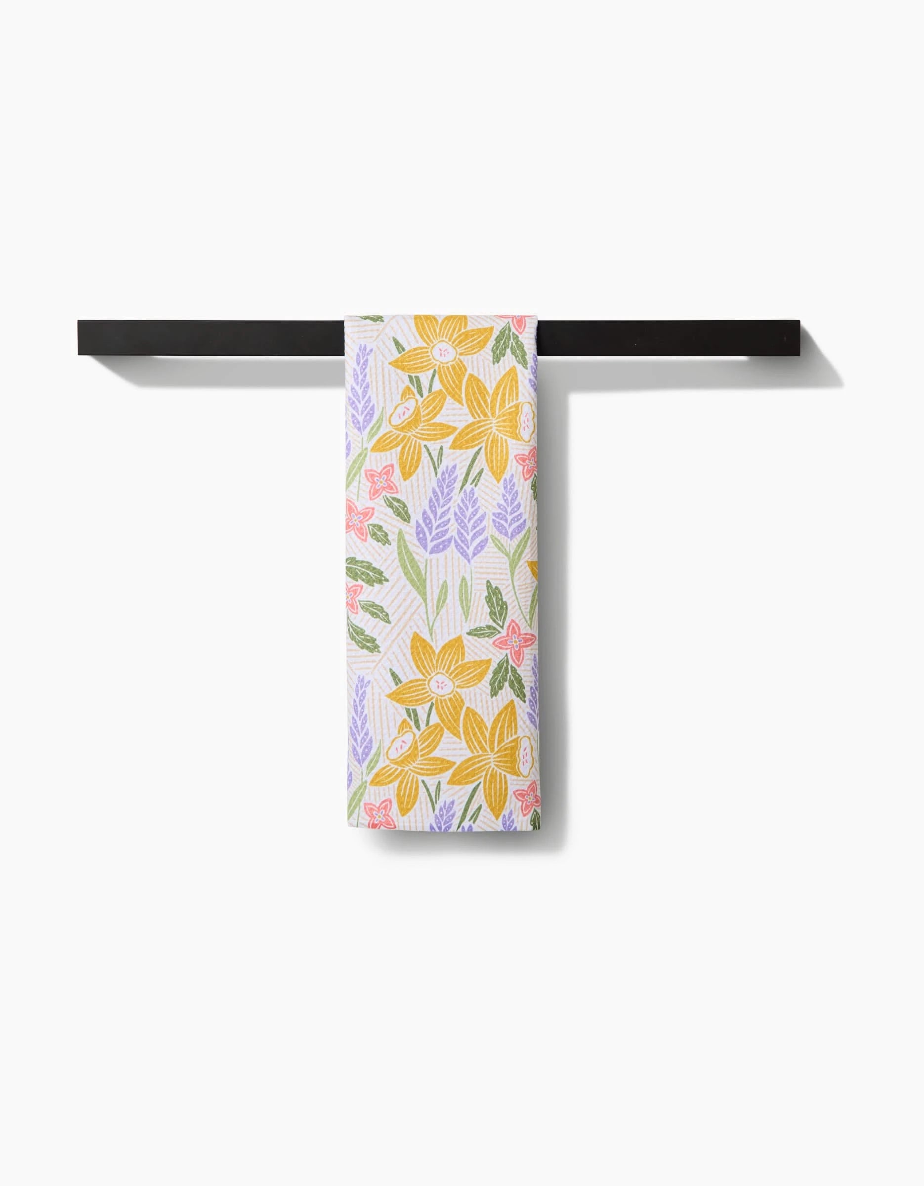 The “Daffodil Dreams” tea towel by Sarah Daugherty features a vibrant floral pattern in yellow, purple, and pink. Made from soft Geoweave fabric, it hangs neatly and adds both style and function to any space.