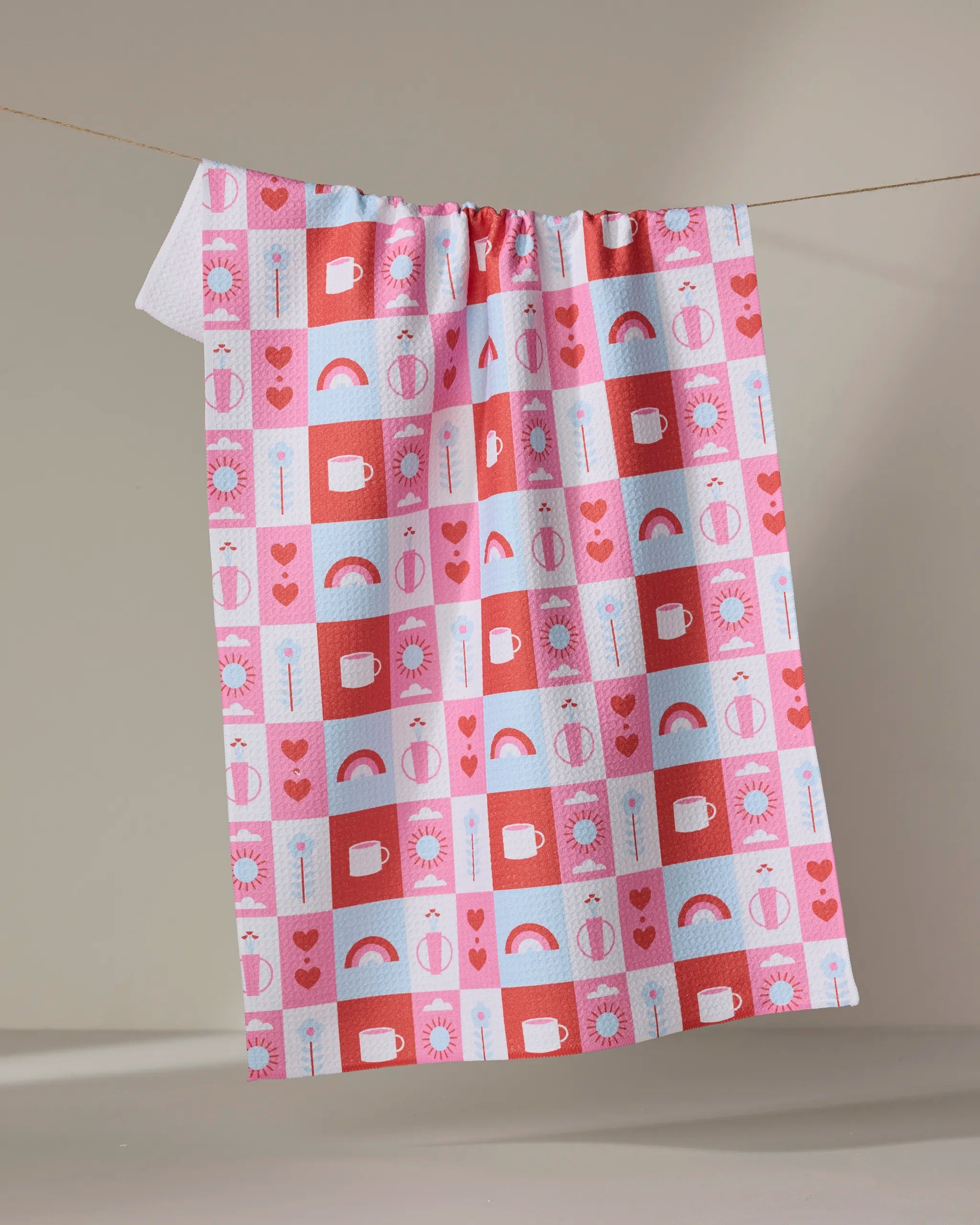 The "Daily Icons" tea towel by Laura Supnik features pink, red, and light blue ultra-absorbent squares adorned with hearts, rainbows, suns, cheese wedges, and arrows, hanging on a string against a plain beige background.