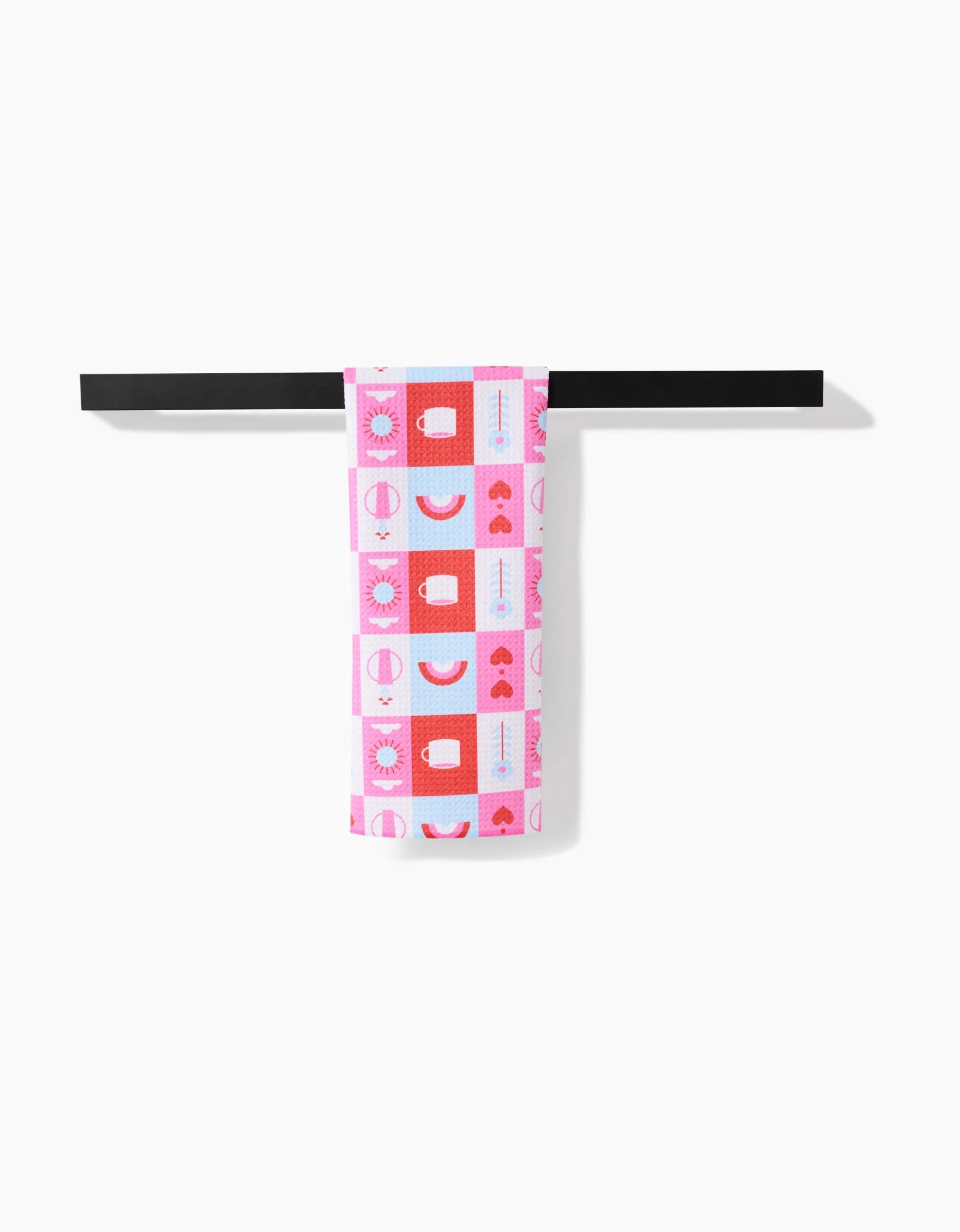 The Daily Icons tea towel by Laura Supnik, made from recycled materials and featuring pink, red, and light blue hearts, circles, and suns, hangs on a simple black towel bar against a white background.