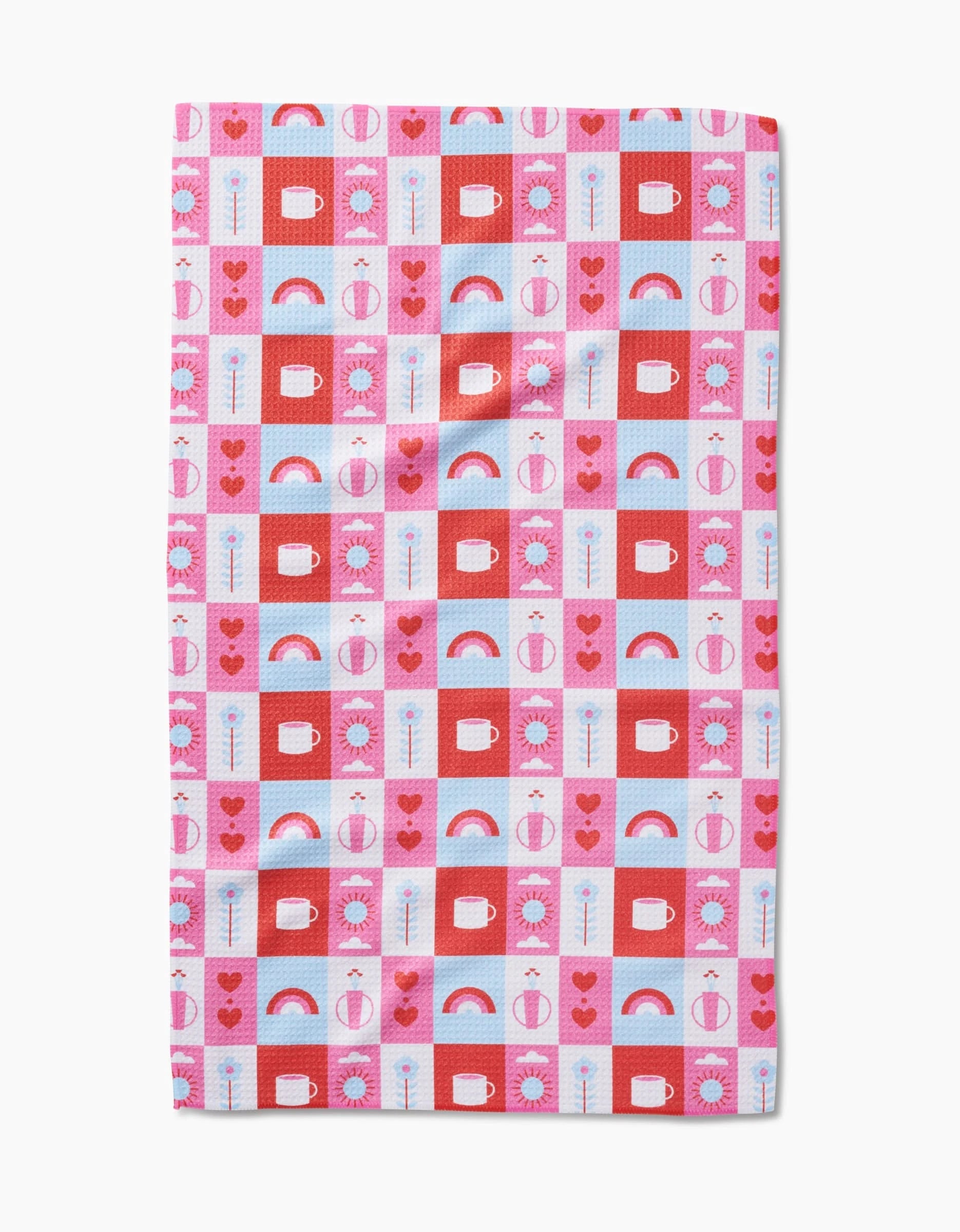 The Daily Icons tea towel by Laura Supnik is rectangular, made from recycled materials, and features a grid of colorful hearts, rainbows, flowers, suns, and abstract shapes in red, pink, blue, and white.