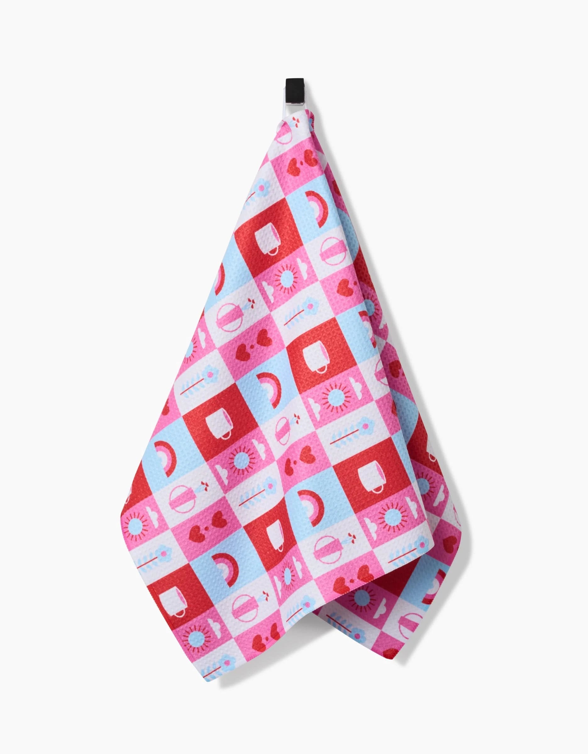 The Daily Icons tea towel by Laura Supnik features pink, red, and blue checks with suns, moons, rainbows, and hearts. Made from recycled materials, it hangs from a black loop against a white background.
