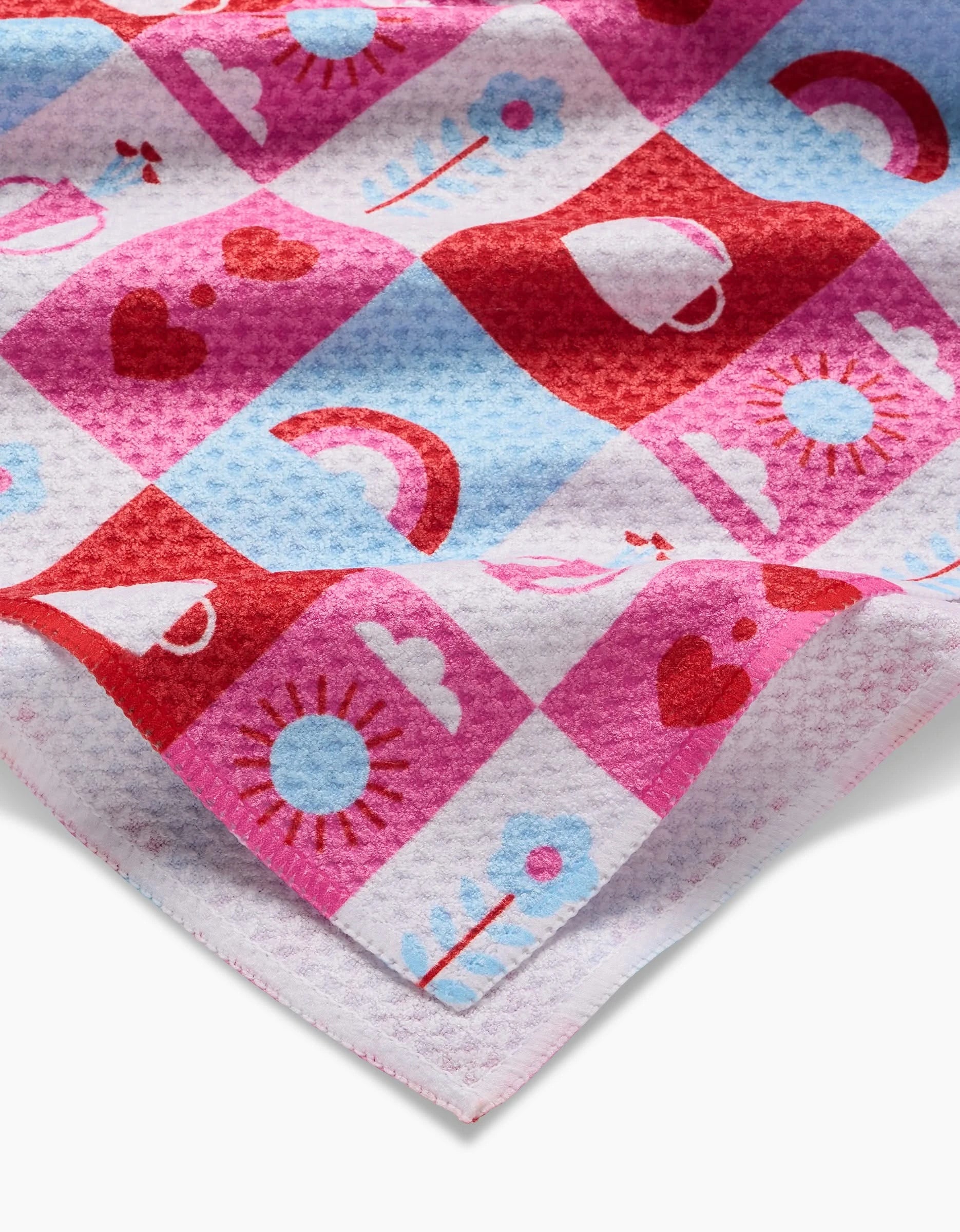 The Laura Supnik Daily Icons tea towel features soft Geoweave fabric with a colorful checkered pattern of hearts, rainbows, suns, flowers, and cups in red, pink, and blue. Made from recycled materials and slightly folded at the corner.