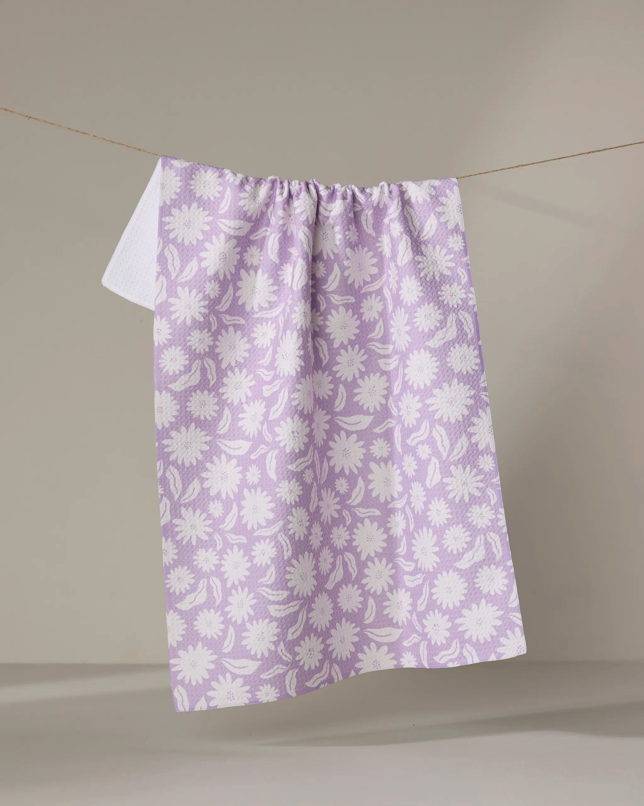 The Kayla Ann Daisy Bloom kitchen tea towel, featuring a lilac and white floral design and made from recycled materials, hangs on a clothesline against a plain light beige background.