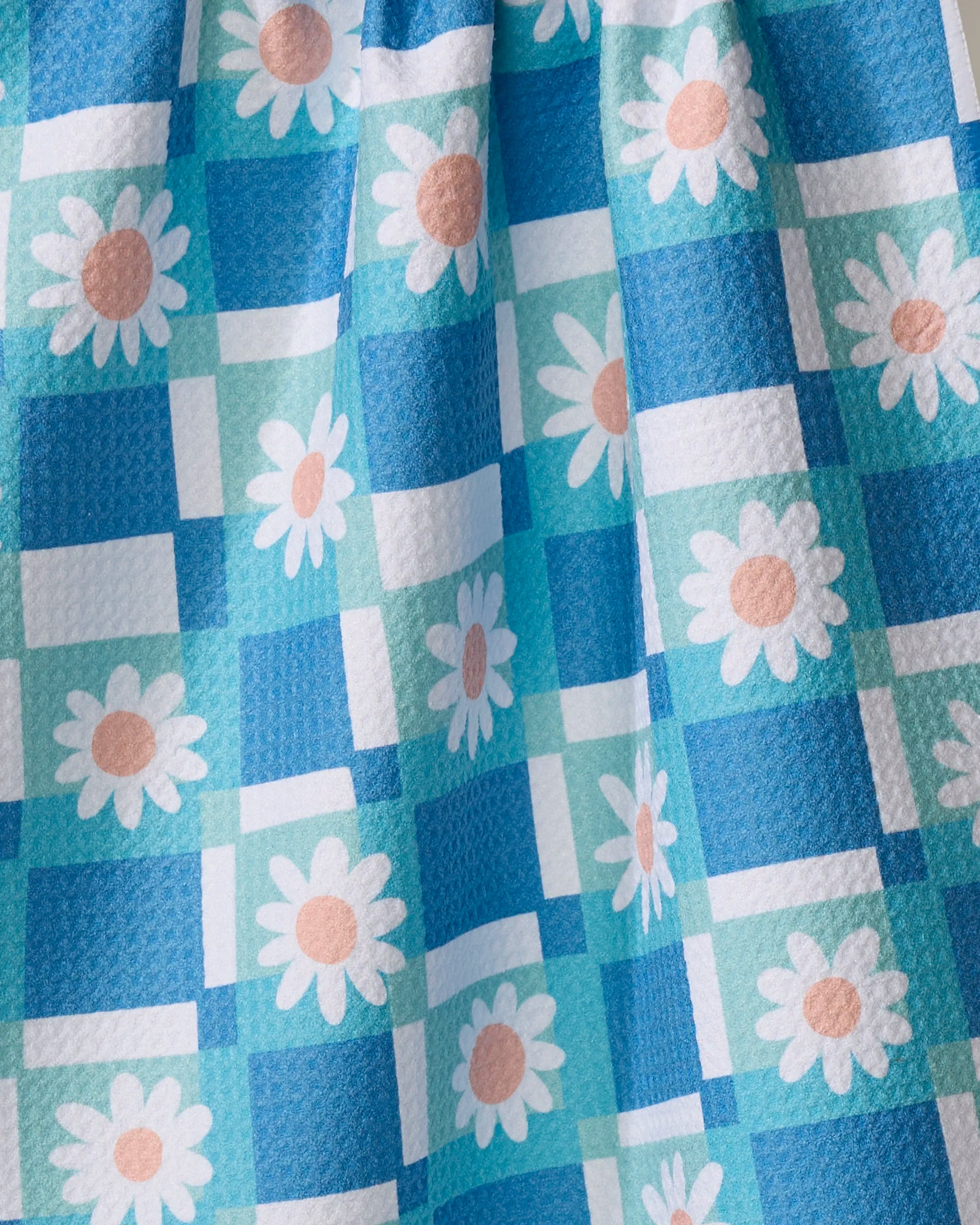 A close-up of Rachel Parker's Daisy Daisy tea towel, featuring a blue and white checkered pattern with white daisies with peach centers, crafted from recycled materials.