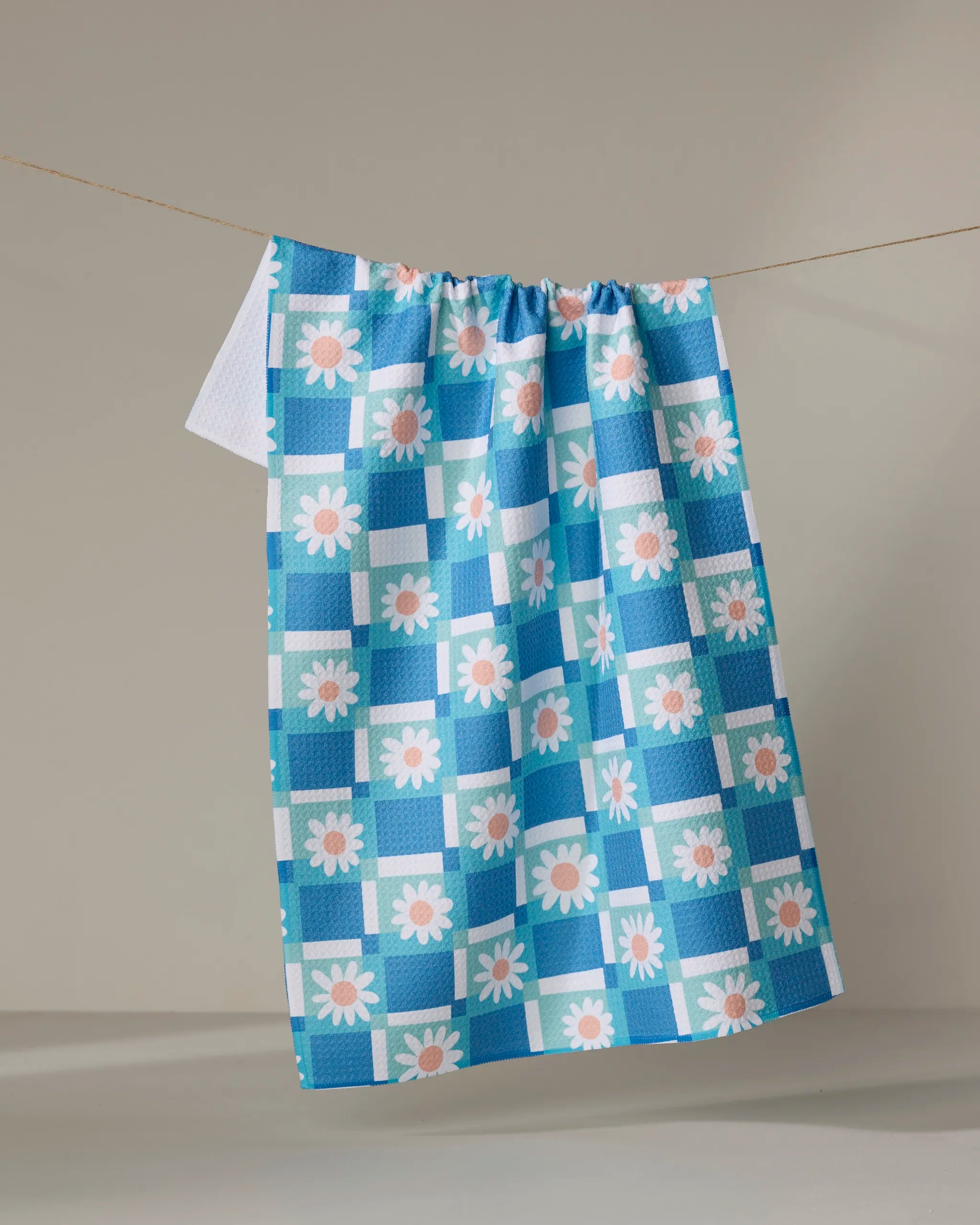 The Daisy Daisy tea towel by Rachel Parker, featuring a blue and white checkered design with daisy florals and made from recycled materials, hangs on a clothesline against a plain light background.