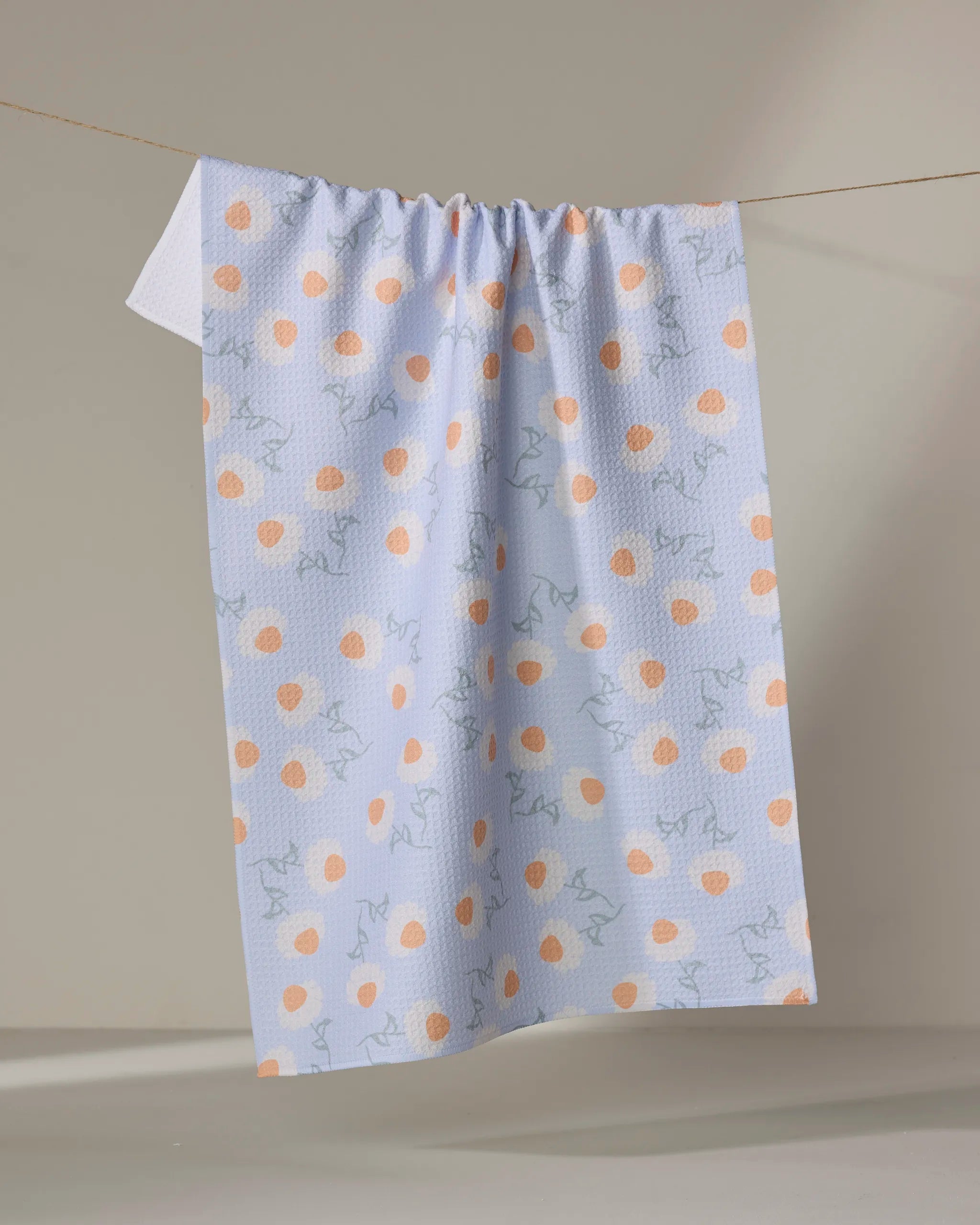The Daisy Duets tea towel by Sarah Reed features orange polka dots and light blue abstract lines on white, made from recycled materials, and hangs on a clothesline against a neutral beige background.