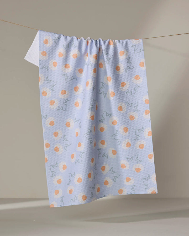 The Daisy Duets tea towel by Sarah Reed features orange polka dots and light blue abstract lines on white, made from recycled materials, and hangs on a clothesline against a neutral beige background.