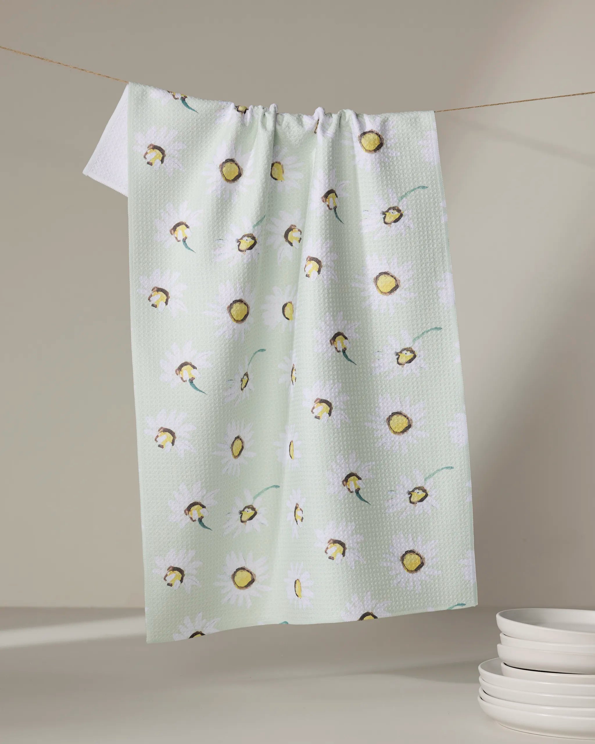 The Daisy Field kitchen towel by Ania Zwara Art, in pale green with a white daisy and yellow center pattern, hangs on a clothesline. White plates are stacked in the bottom right corner against a neutral background.