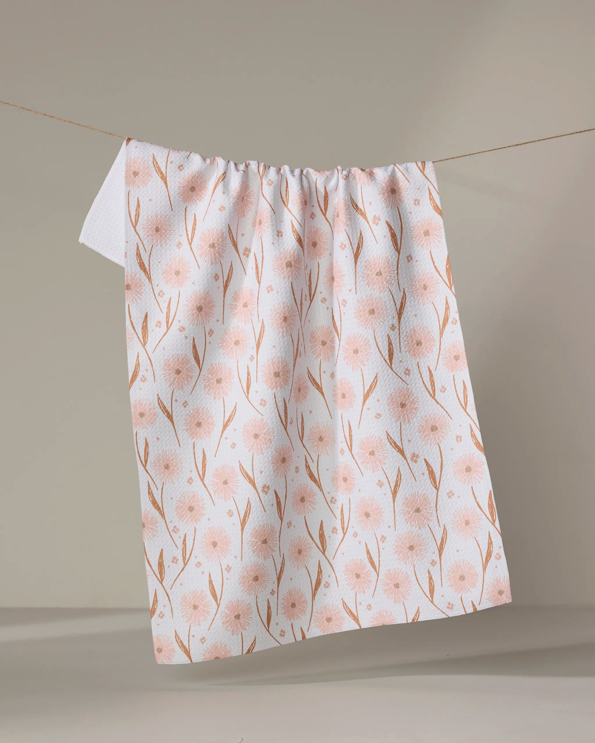 The Daisy Pop tea towel by Hufton Studio, featuring a light pink floral design on white, hangs on a clothesline against a neutral background. Its quick-drying fabric makes it both stylish and practical for any kitchen.