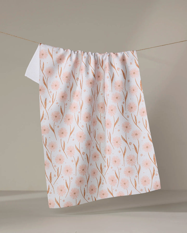The Daisy Pop tea towel by Hufton Studio, featuring a light pink floral design on white, hangs on a clothesline against a neutral background. Its quick-drying fabric makes it both stylish and practical for any kitchen.