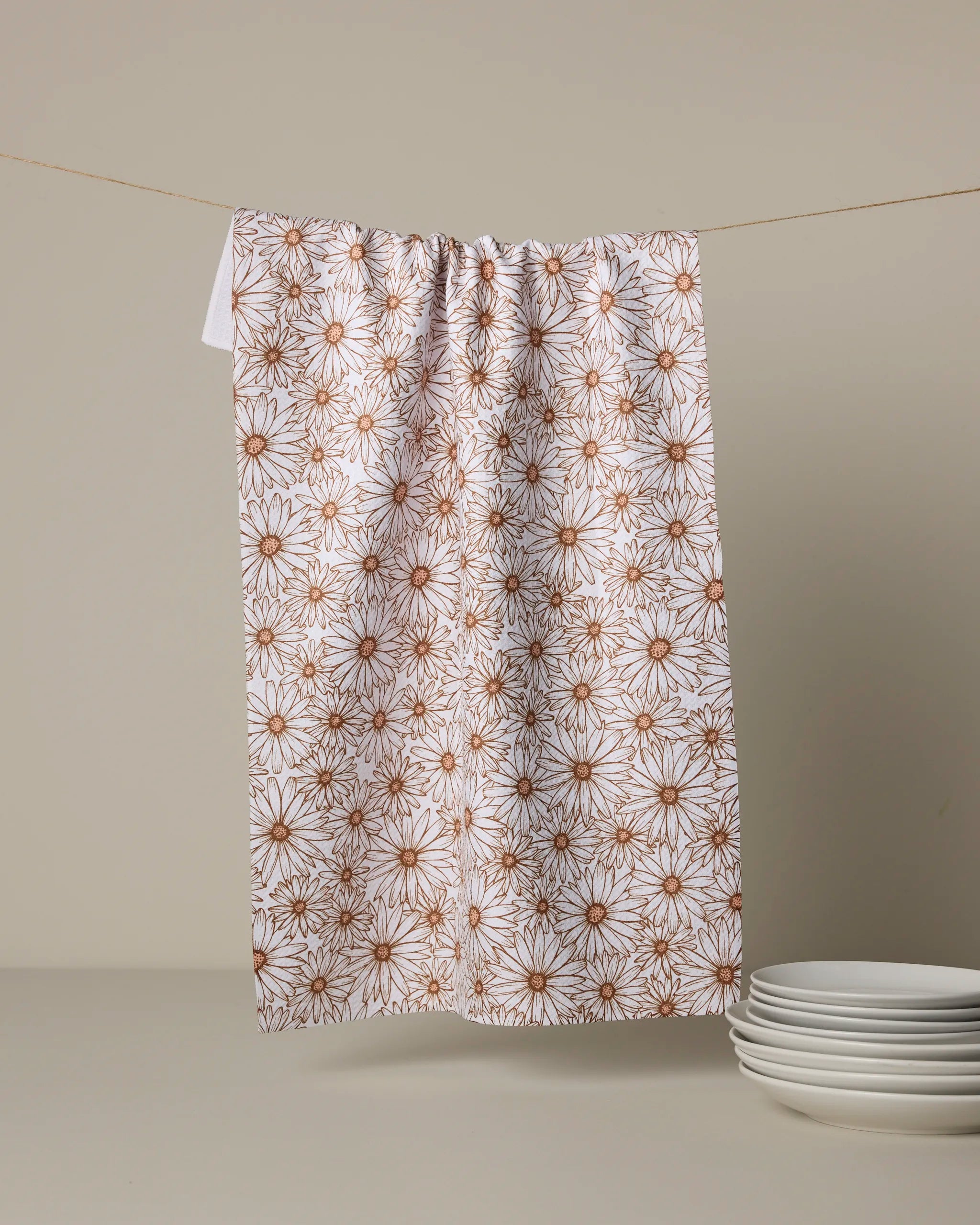 Geometry's Daisy Trail tea towel, ultra-absorbent with a brown daisy pattern on white, hangs beside four stacked white plates against a beige wall.