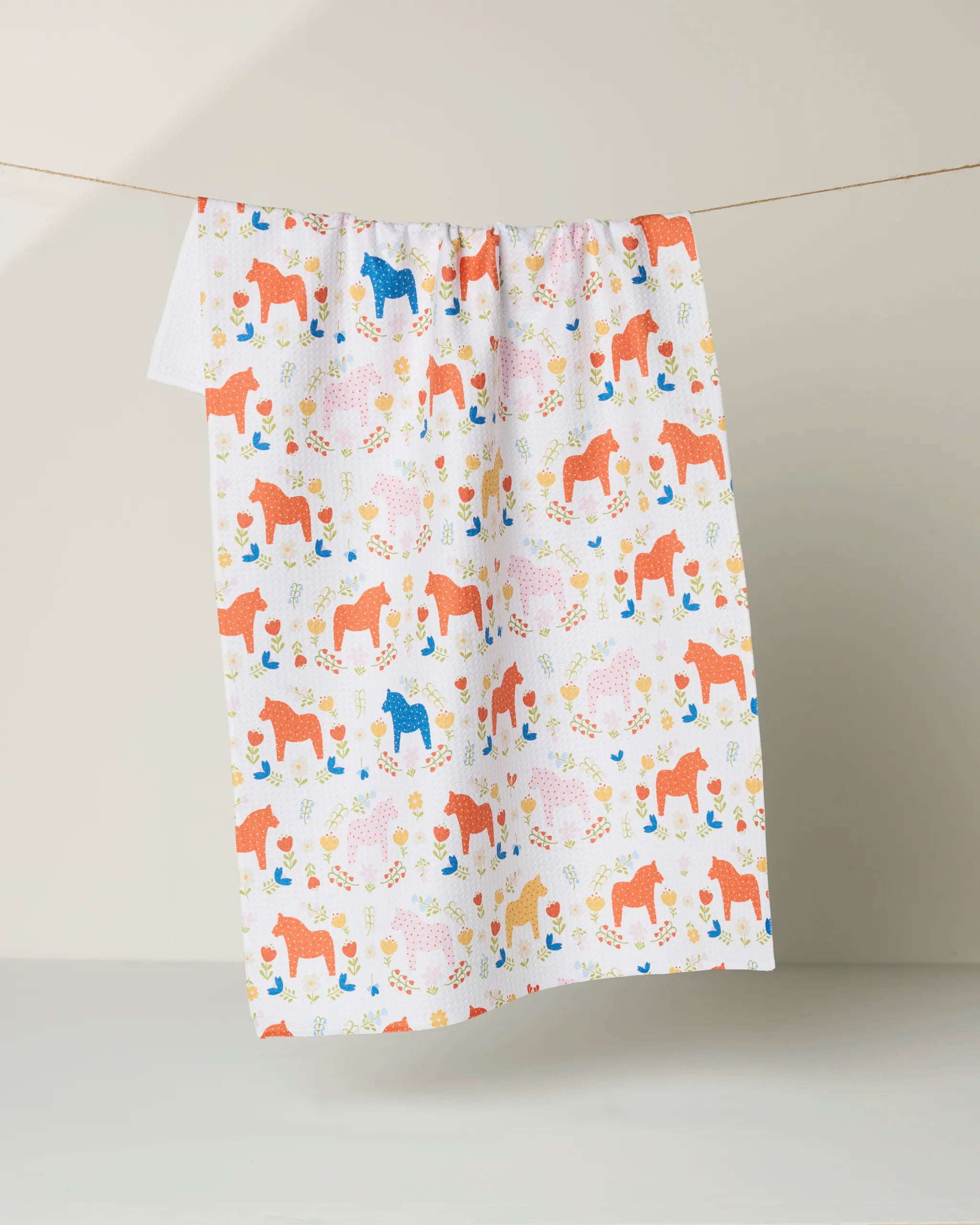 The Sara Mari Dala Garden Floral kitchen towel, made from recycled materials and adorned with colorful red, blue, pink, and yellow horses, flowers, and leaves, hangs on a line against a neutral background.