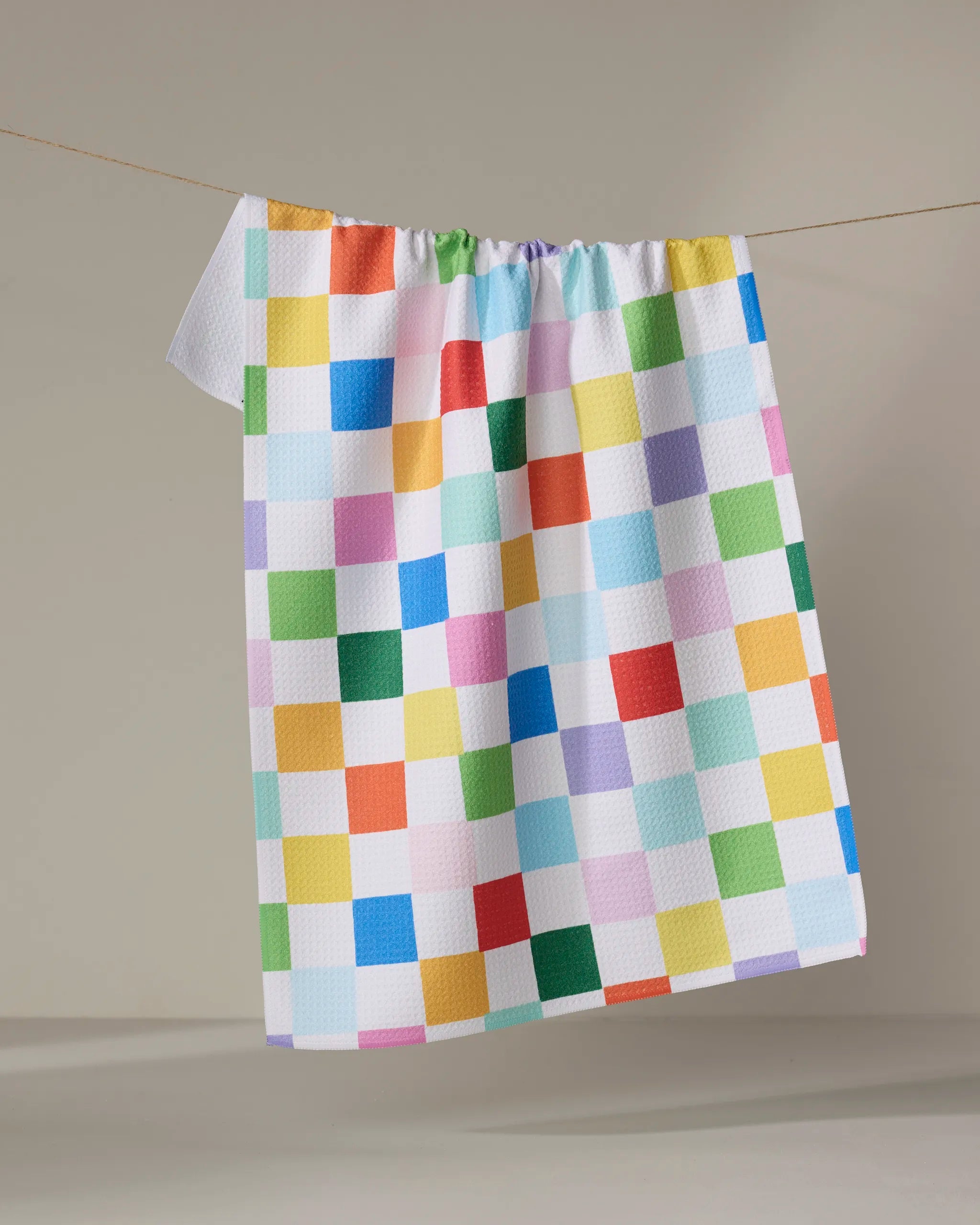 The Dance Floor dish towel by Vaishali Design Studio features a colorful checkered pattern in blue, green, yellow, orange, red, and pink on white and hangs on a clothesline against a plain beige background.