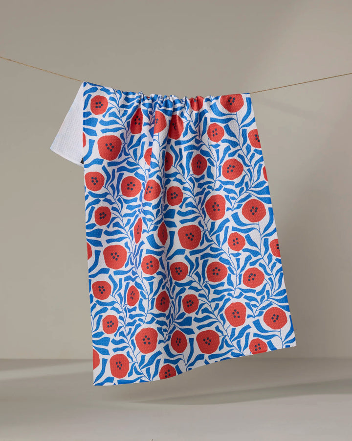 The Dancing Poppies tea towel by Happy Hemera, made from recycled materials, displays vibrant red flowers and blue leaves and hangs on a clothesline against a neutral background.