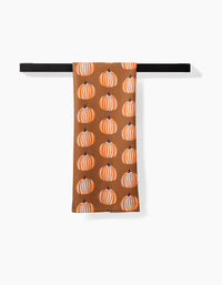 The Lizzie Clark Dancing Pumpkins towel—a brown, ultra-absorbent towel featuring orange and white pumpkin patterns—hangs over a black towel bar against a white background.