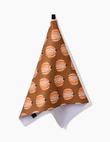 The “Dancing Pumpkins” towel by Lizzie Clark features an ultra-absorbent brown fabric, decorated with orange and white striped pumpkins, and hangs from a black loop on a plain white background.