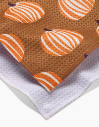 A close-up of two overlapping towels: on top is Lizzie Clark’s Dancing Pumpkins tea towel, brown with orange and white pumpkin patterns; beneath is a white recycled kitchen towel with a textured, waffle-like surface.