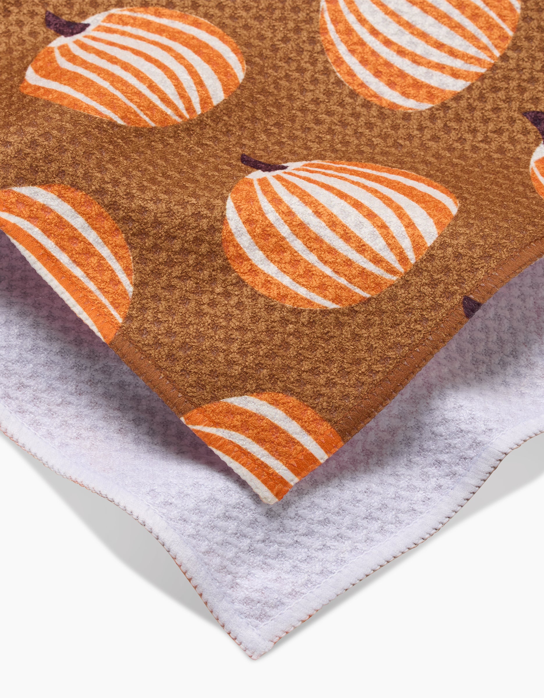 A close-up of two overlapping towels: on top is Lizzie Clark’s Dancing Pumpkins tea towel, brown with orange and white pumpkin patterns; beneath is a white recycled kitchen towel with a textured, waffle-like surface.