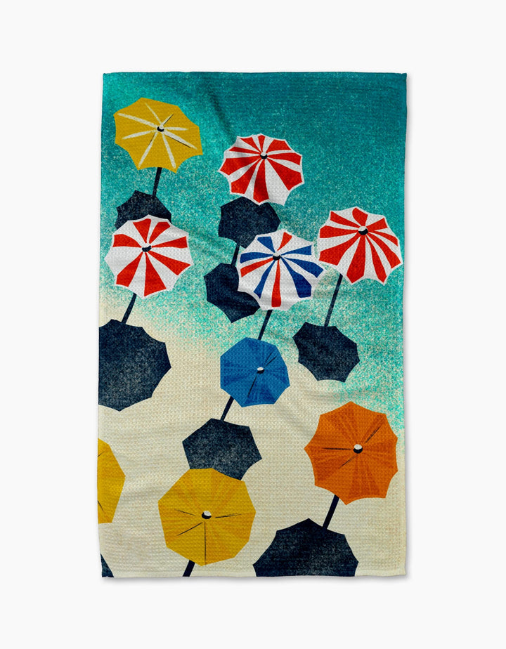The Dancing Umbrellas beach towel by Geometry is made from ultra-absorbent Geoweave fabric and showcases an aerial design with colorful umbrellas in yellow, blue, black, red, and white on sand against a teal sea backdrop.