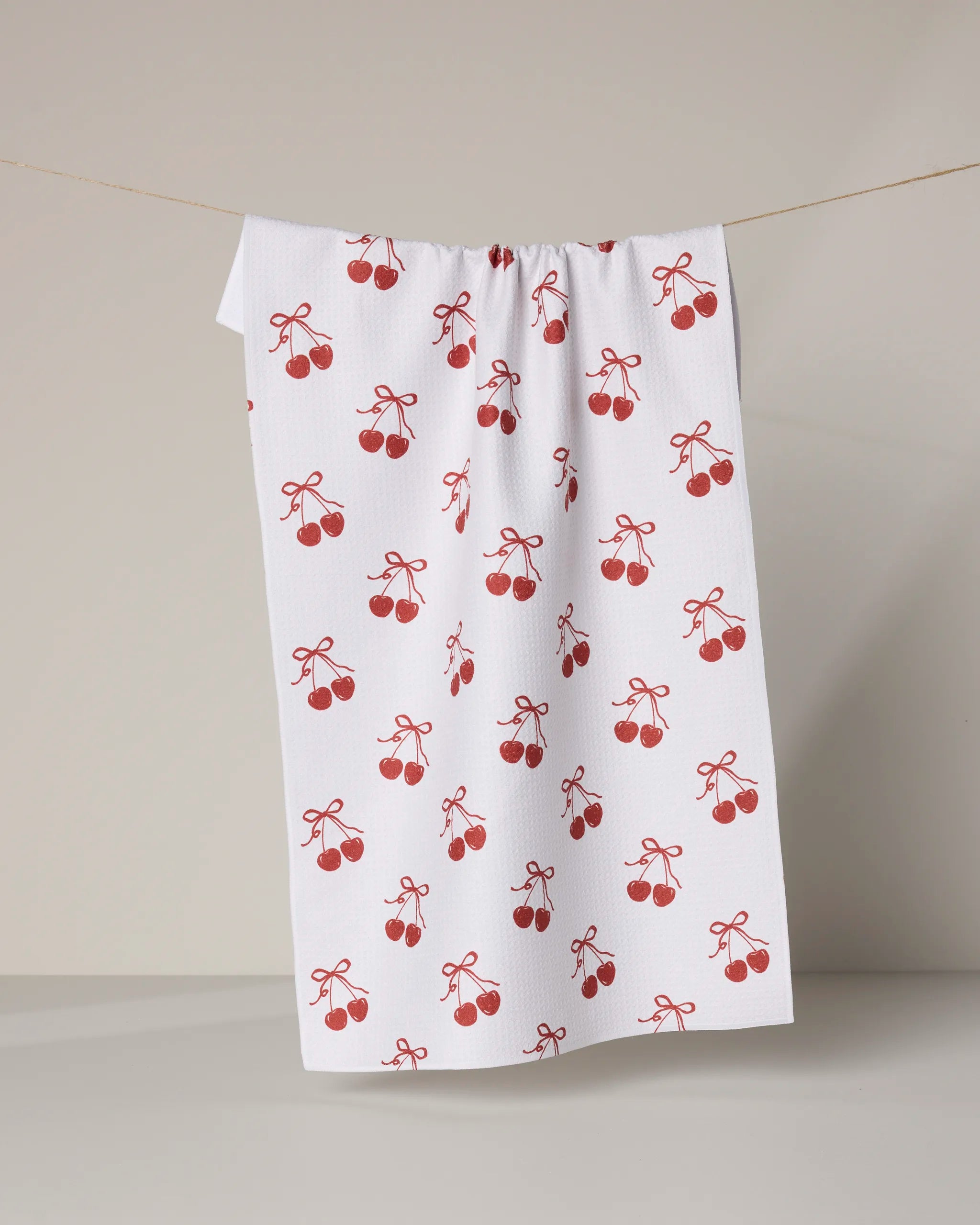 The Geometry "Darling" towel, ultra-absorbent and white, features a repeating pattern of red cherries tied with red bows. It hangs on a line against a clean, neutral background.