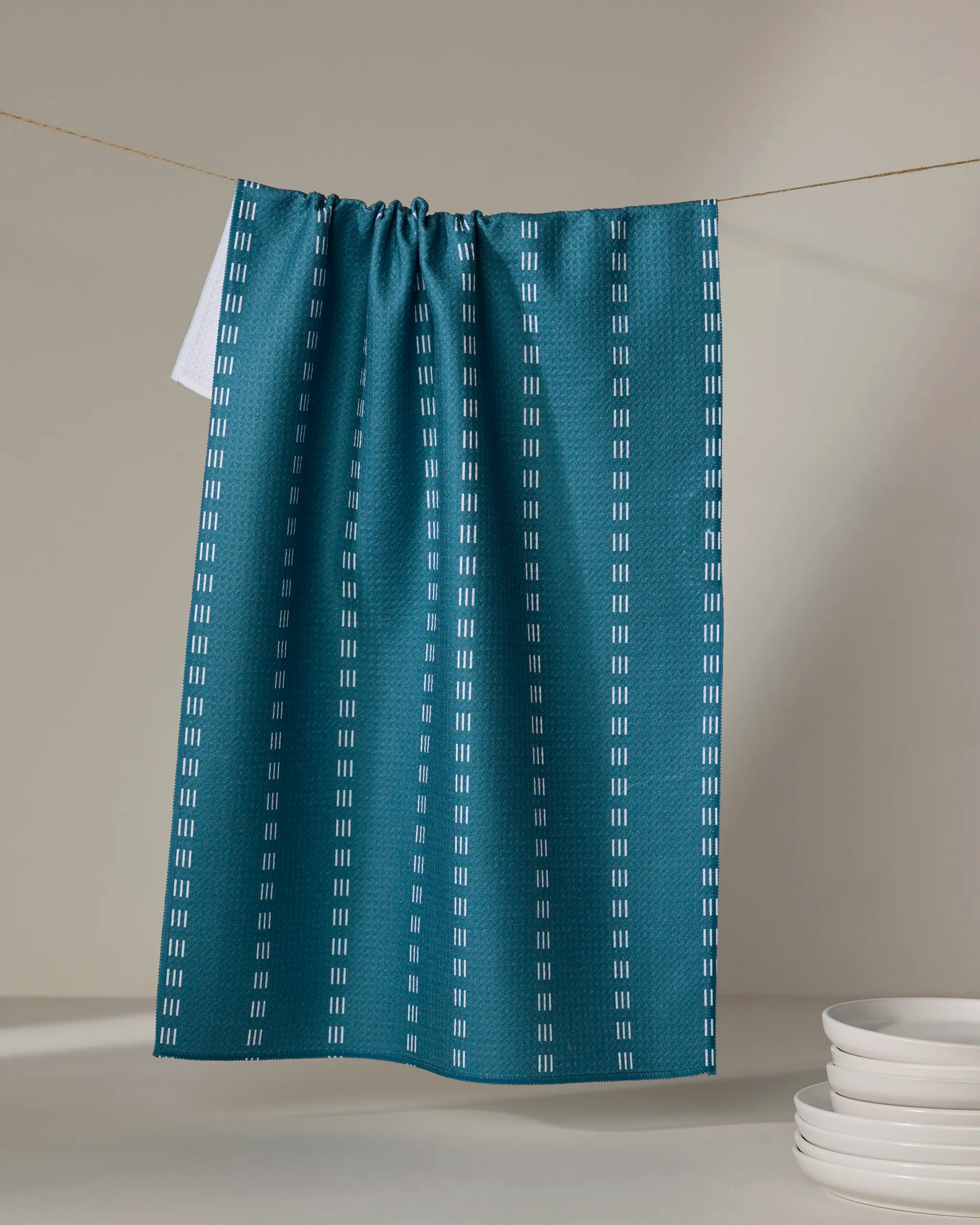 The Buy Guide’s Dashing - Emerald tea towel, crafted from recycled materials with vertical white dashed lines, hangs on a string. White plates are stacked in the bottom right corner against a neutral background.