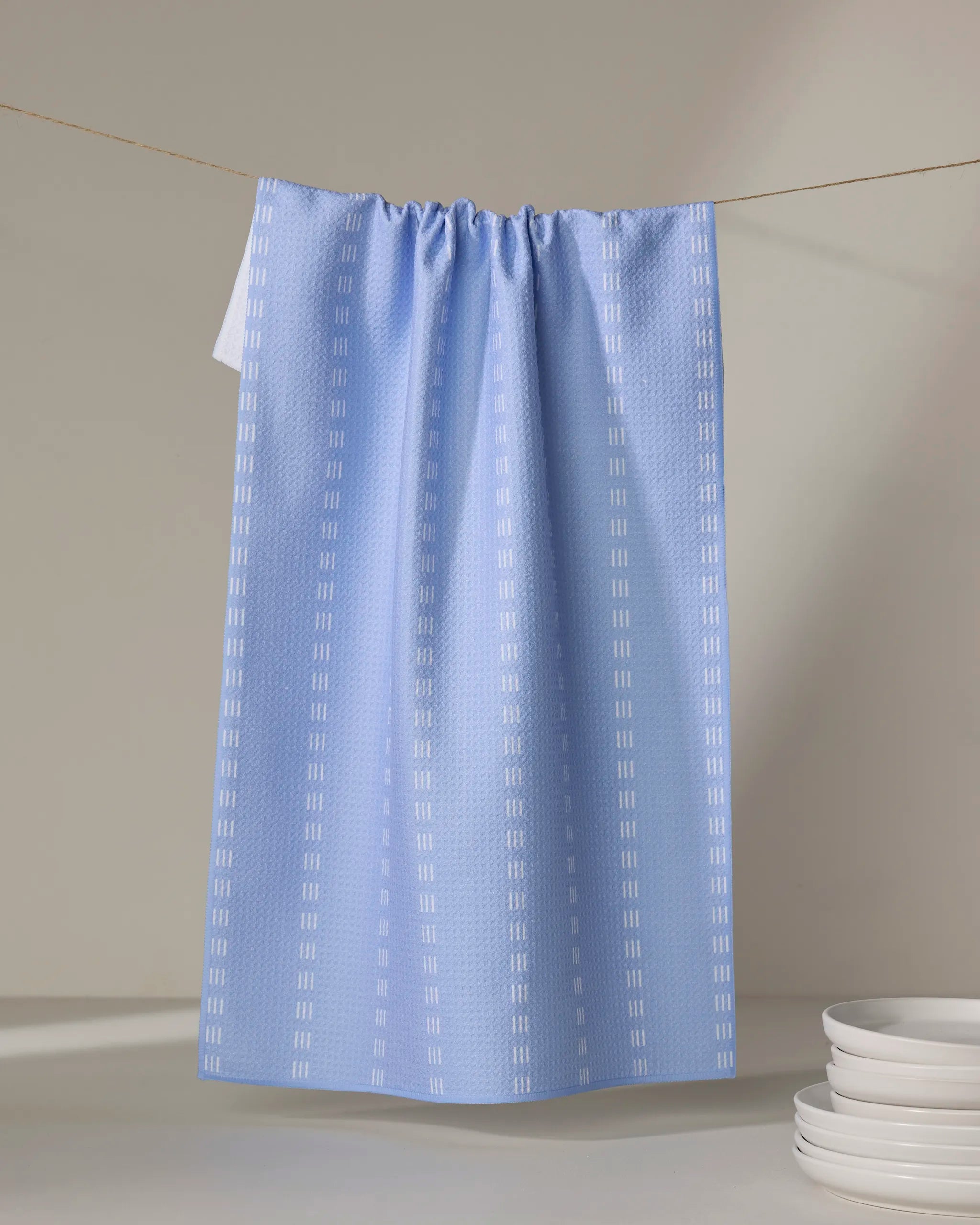 A Dashing - French Blue kitchen towel by The Buy Guide, featuring vertical white dashed lines, hangs on a clothesline near a stack of white plates. Made from recycled materials, this tea towel stands out against a neutral background.