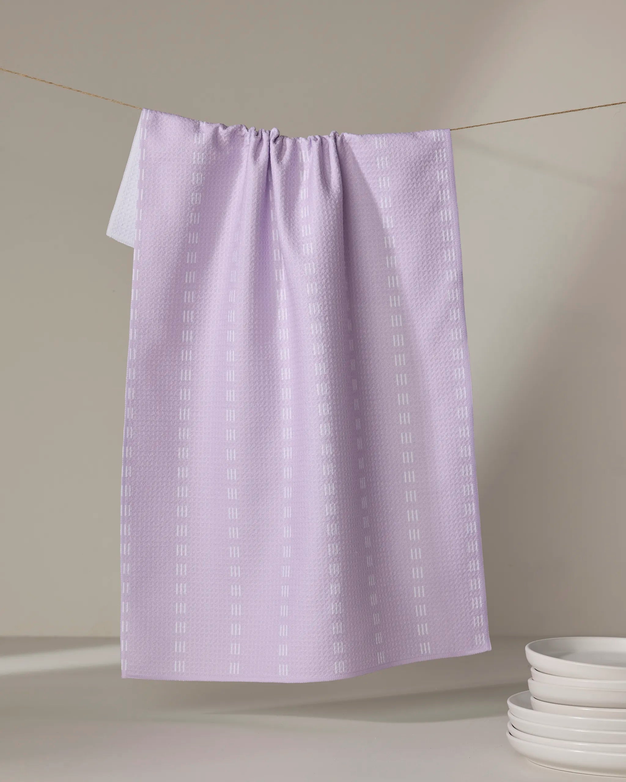 The Buy Guide's Dashing - Lilac tea towel, crafted from recycled materials, hangs on a clothesline showcasing its textured pattern. Nearby, white plates are stacked neatly on a light surface against a softly lit, neutral backdrop.