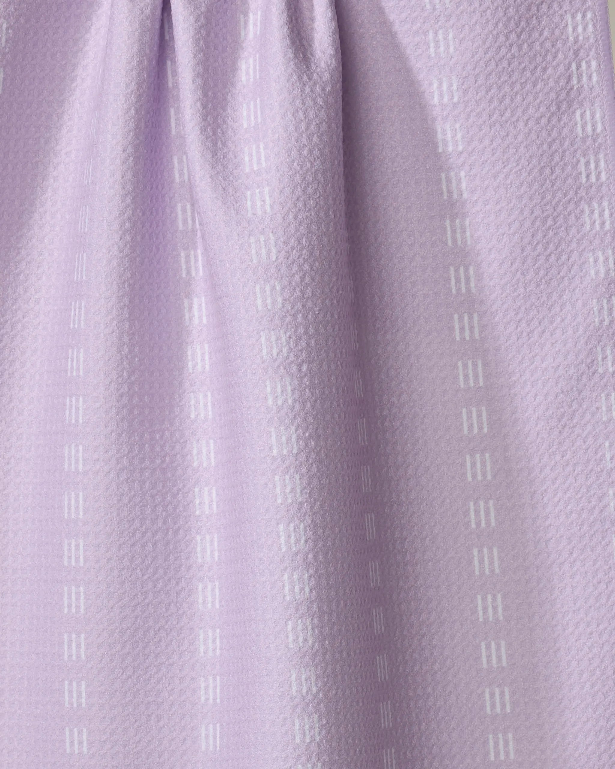 Dashing - Lilac by The Buy Guide is a light purple tea towel featuring a subtle textured pattern with vertical rows of small white rectangles. Made from recycled materials, its fabric is softly gathered at the top for gentle folds.