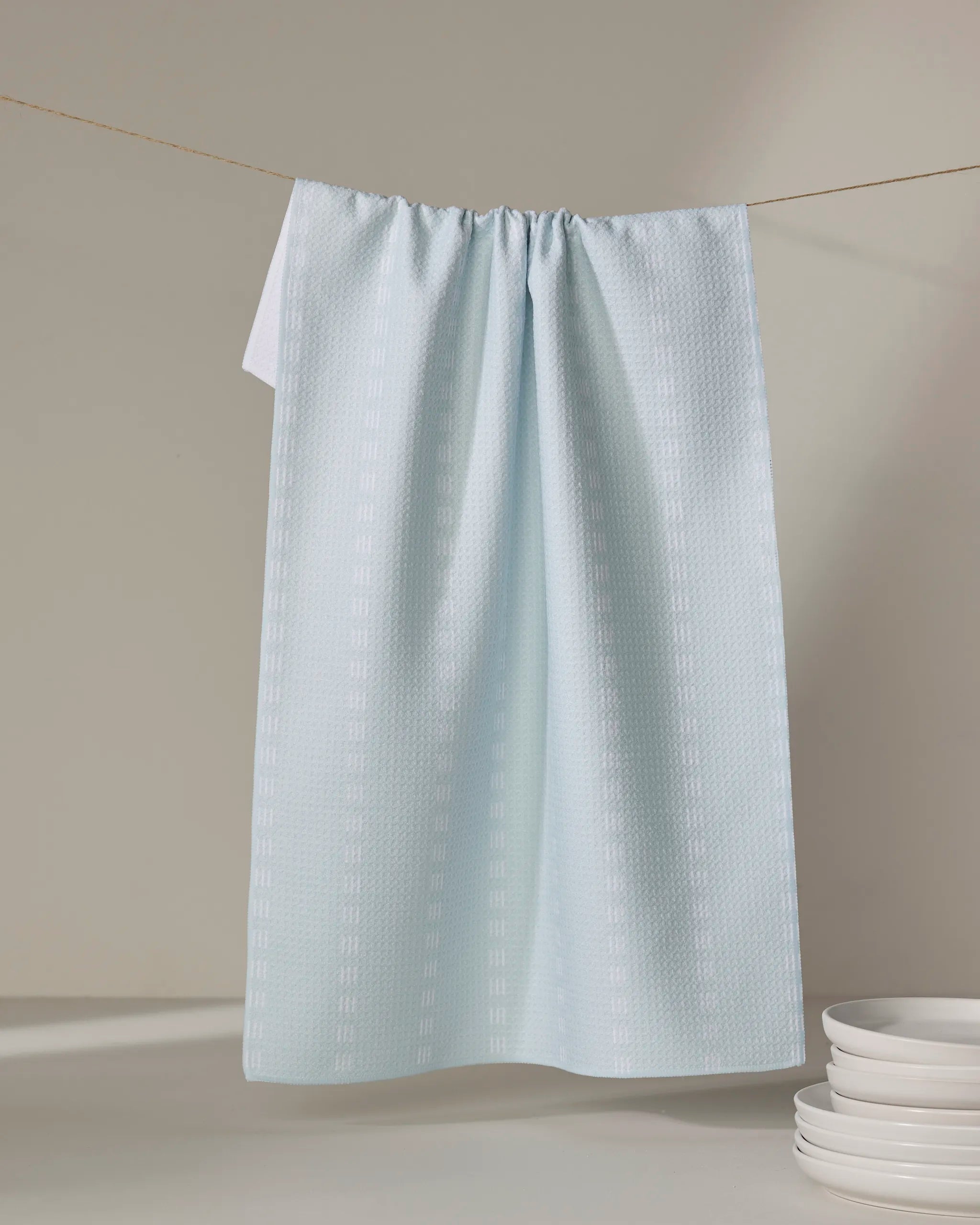 A Dashing - Mint tea towel from The Buy Guide, made of quick-drying recycled materials, hangs on a string against a neutral background with white plates stacked in the lower right corner.