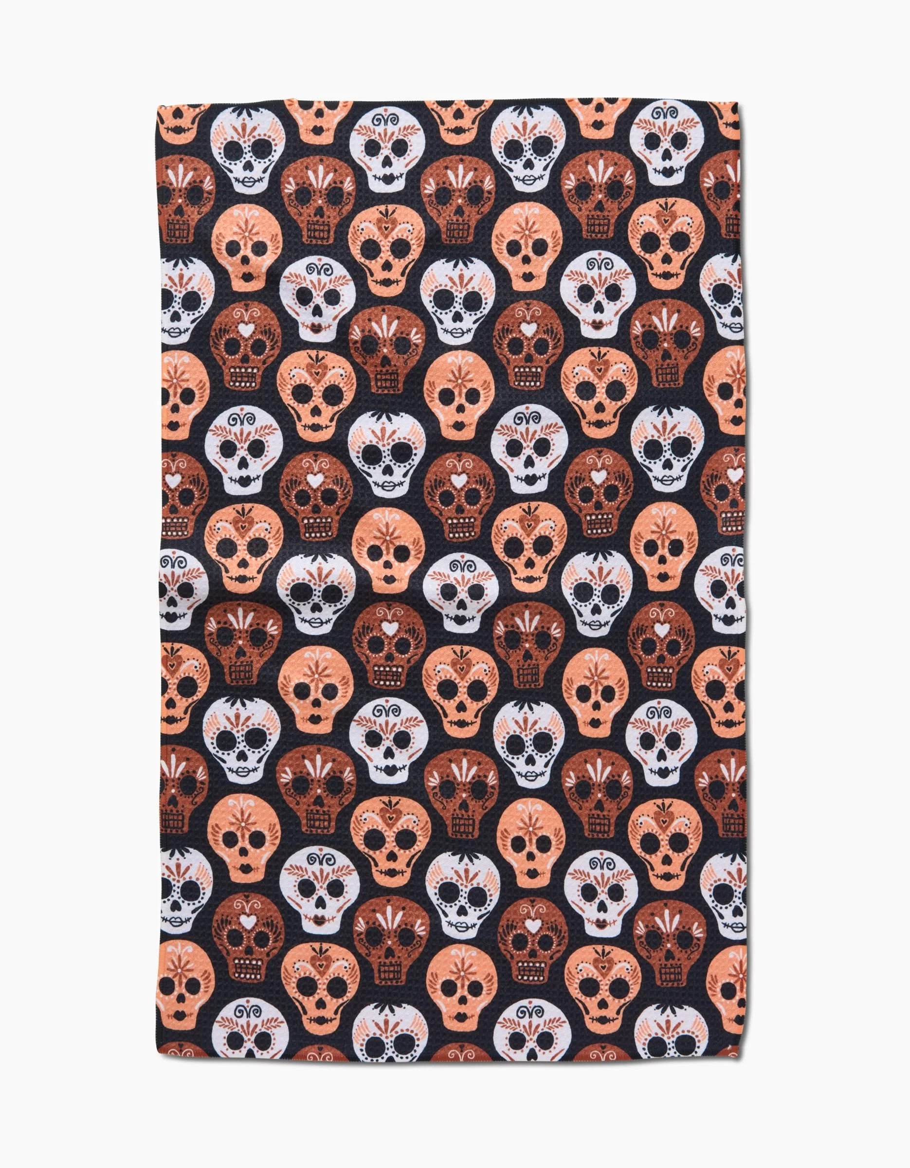 The Day Of The Dead Skulls tea towel by Hufton Studio is rectangular, made from recycled materials, and features a dense pattern of orange, white, and black Día de los Muertos skulls on a dark background.
