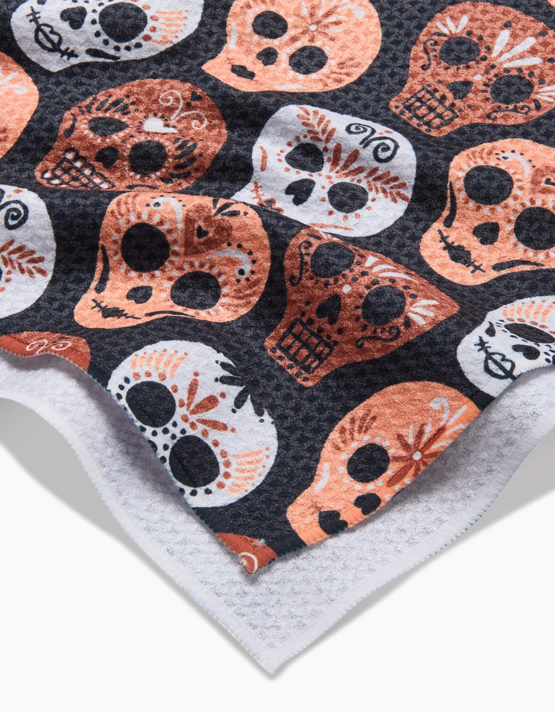 A close-up of the Hufton Studio Day Of The Dead Skulls quick-drying tea towel, made from recycled materials, with orange, white, and black skulls on a dark background. The fabric’s edge appears in the corner.