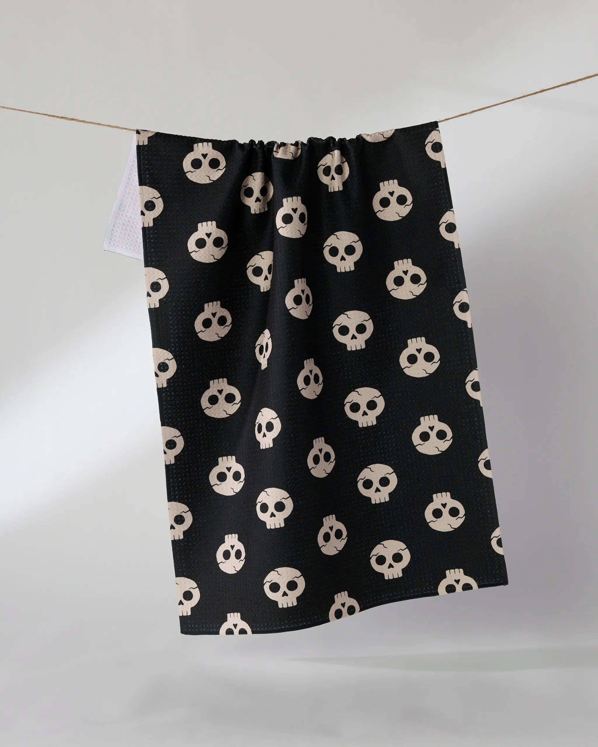 Geometry’s “Dead Cute” tea towel, ultra-absorbent and black with a playful light beige cartoon skull pattern, hangs on a clothesline against a plain light gray background.
