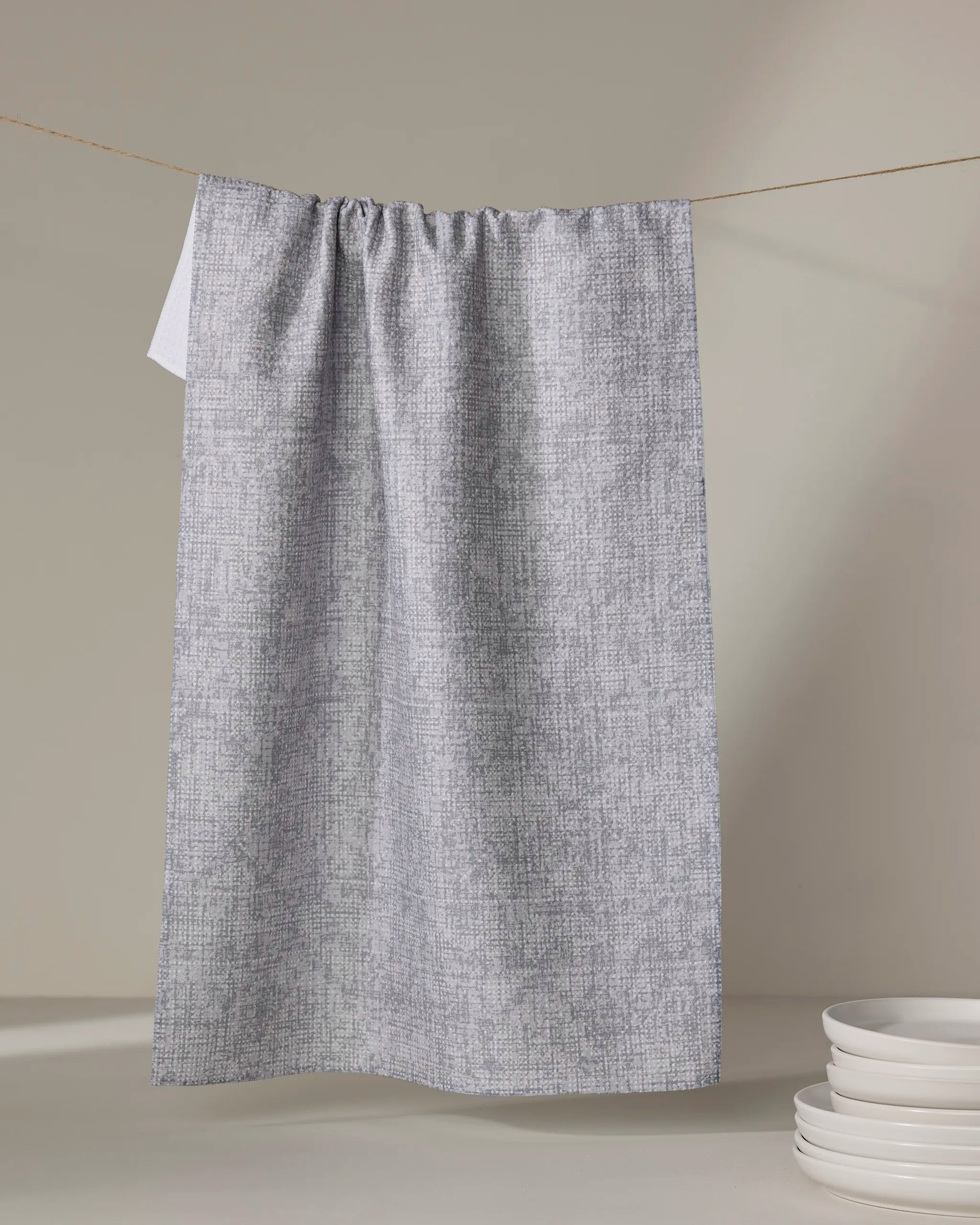 The Debra Grey kitchen towel by Becki Owens hangs from a string against a neutral background, with a stack of white plates placed nearby.