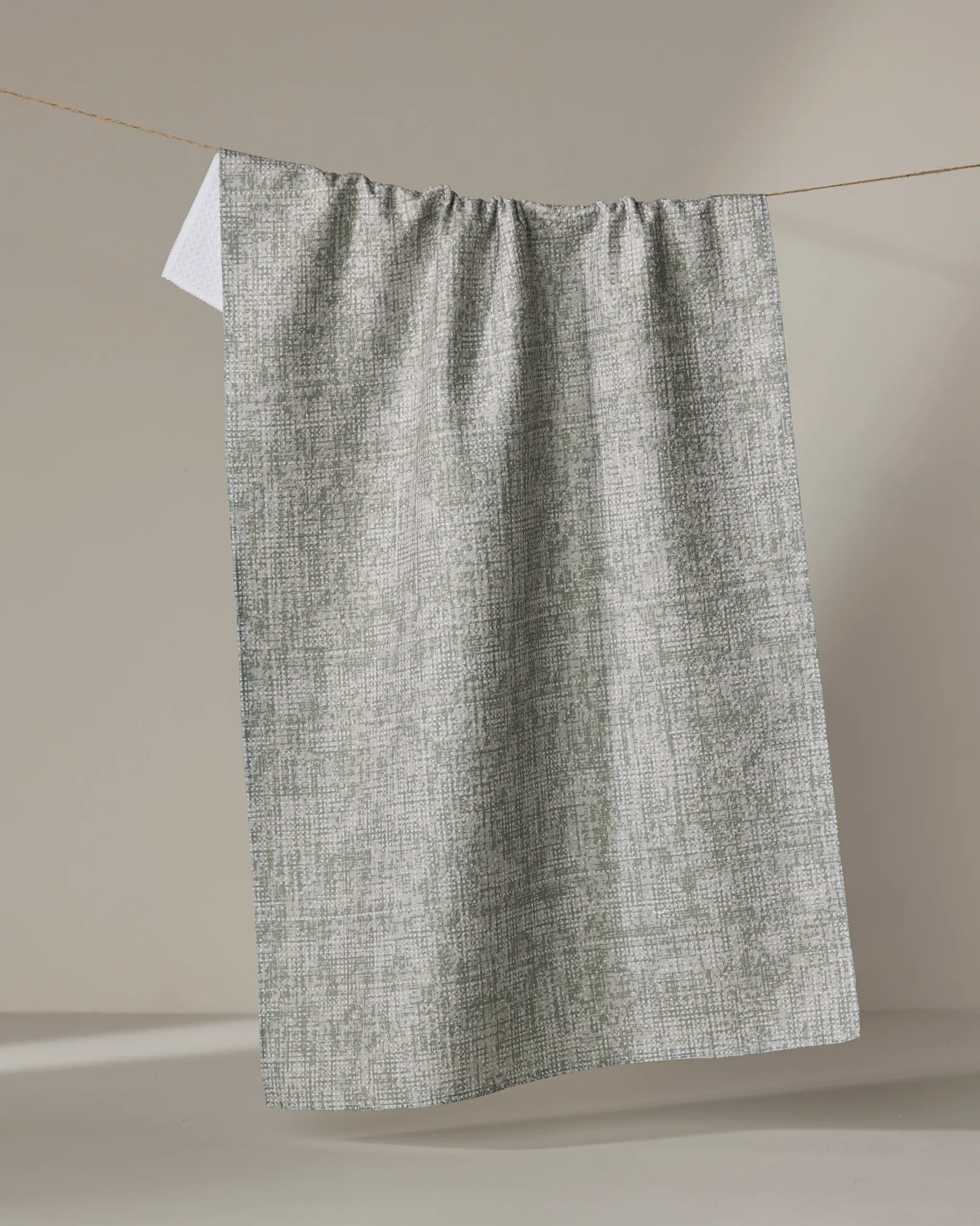 The Debra Sage tea towel by Becki Owens, featuring a gray and white textured design made from recycled materials, hangs neatly on a clothesline against a plain beige background.