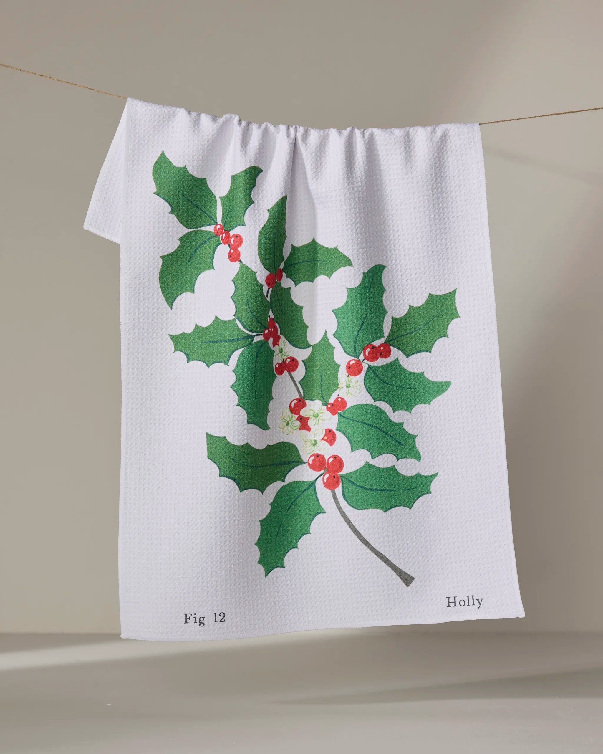 The December Holly tea towel by Linda Fox is made from recycled materials and features a holly branch with green leaves, red berries, and the words "Fig 12" and "Holly" printed at the bottom.