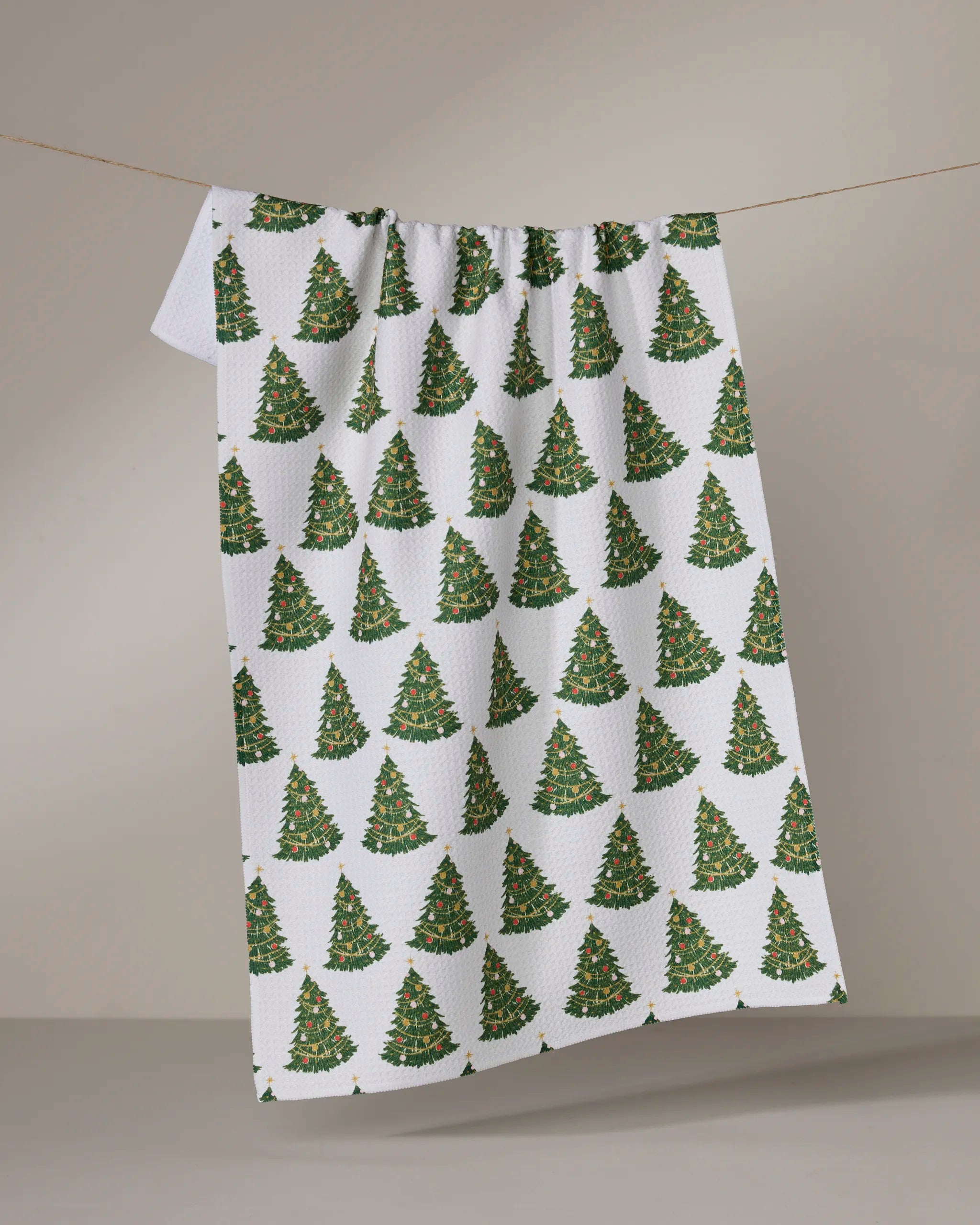 The Geometry "Decked Out" tea towel, made from recycled materials, displays a pattern of green Christmas trees and hangs against a plain neutral background.