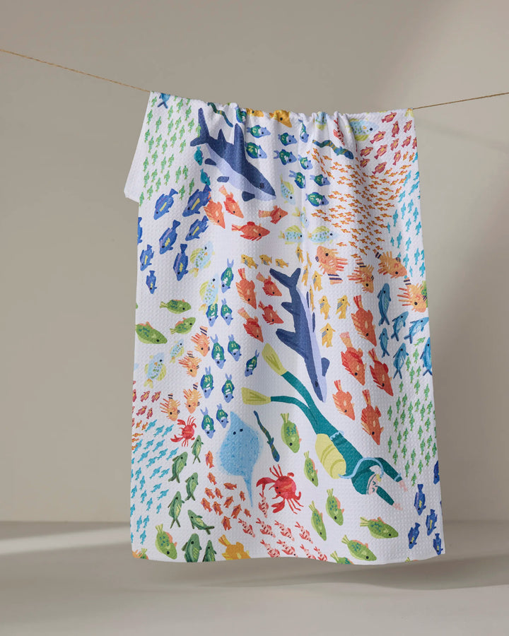 The "Deep Sea Encounter" tea towel by Janna Sue Design features vibrant sharks, fish, crabs, and seaweed, sustainably crafted from recycled materials and shown hanging on a clothesline against a simple backdrop.