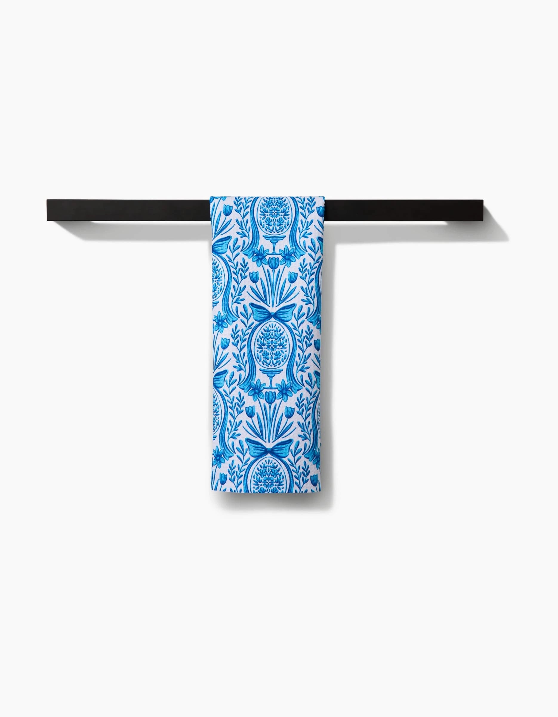 The Sarah Daugherty Delft Blue Easter Egg Damask tea towel, featuring a blue and white floral and tree pattern, hangs neatly over a simple black towel bar against a white background.