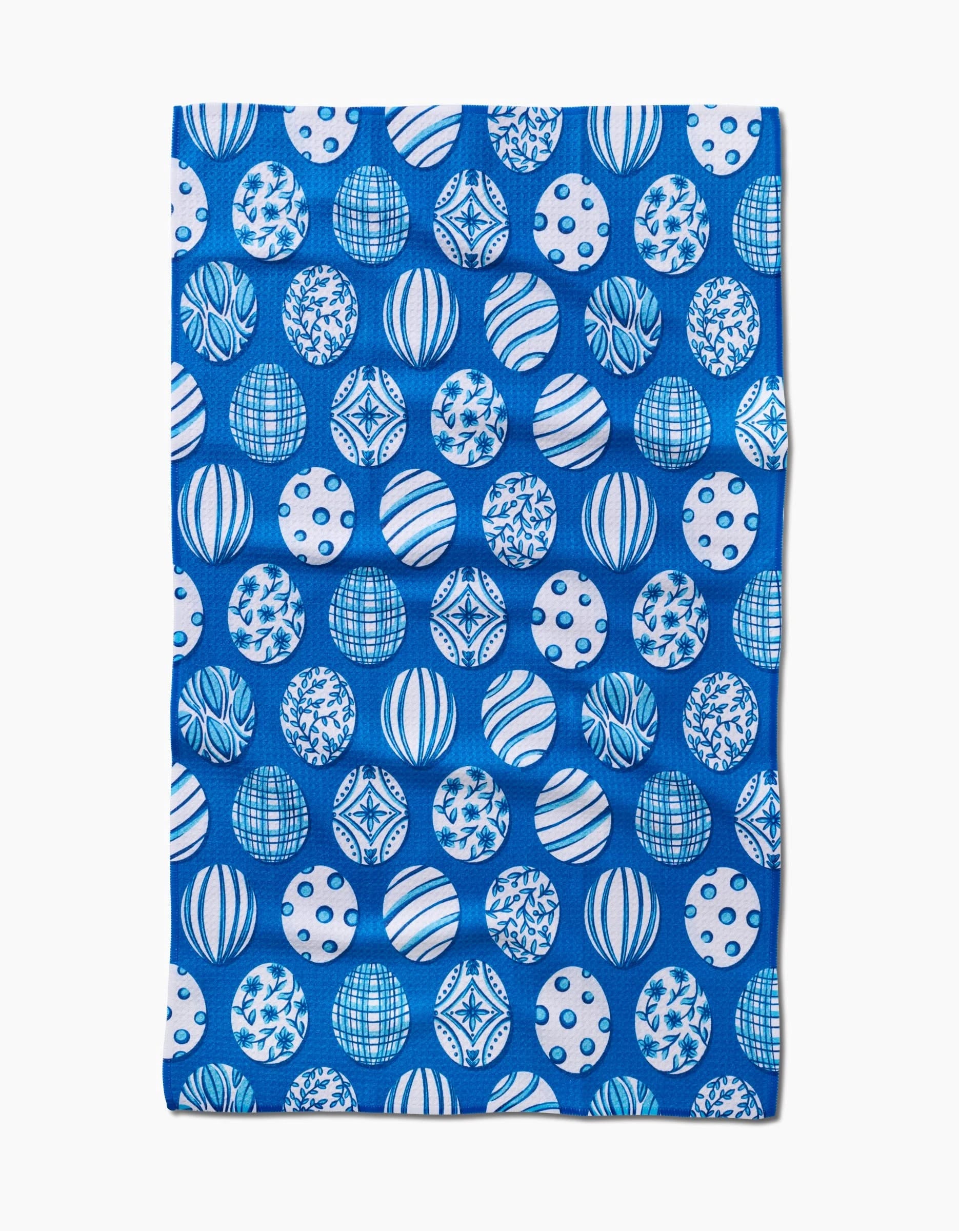 The Delft Blue Easter Eggs tea towel by Sarah Daugherty is rectangular, blue, and made from recycled materials, featuring a repeating pattern of white and blue spheres with geometric and abstract designs.