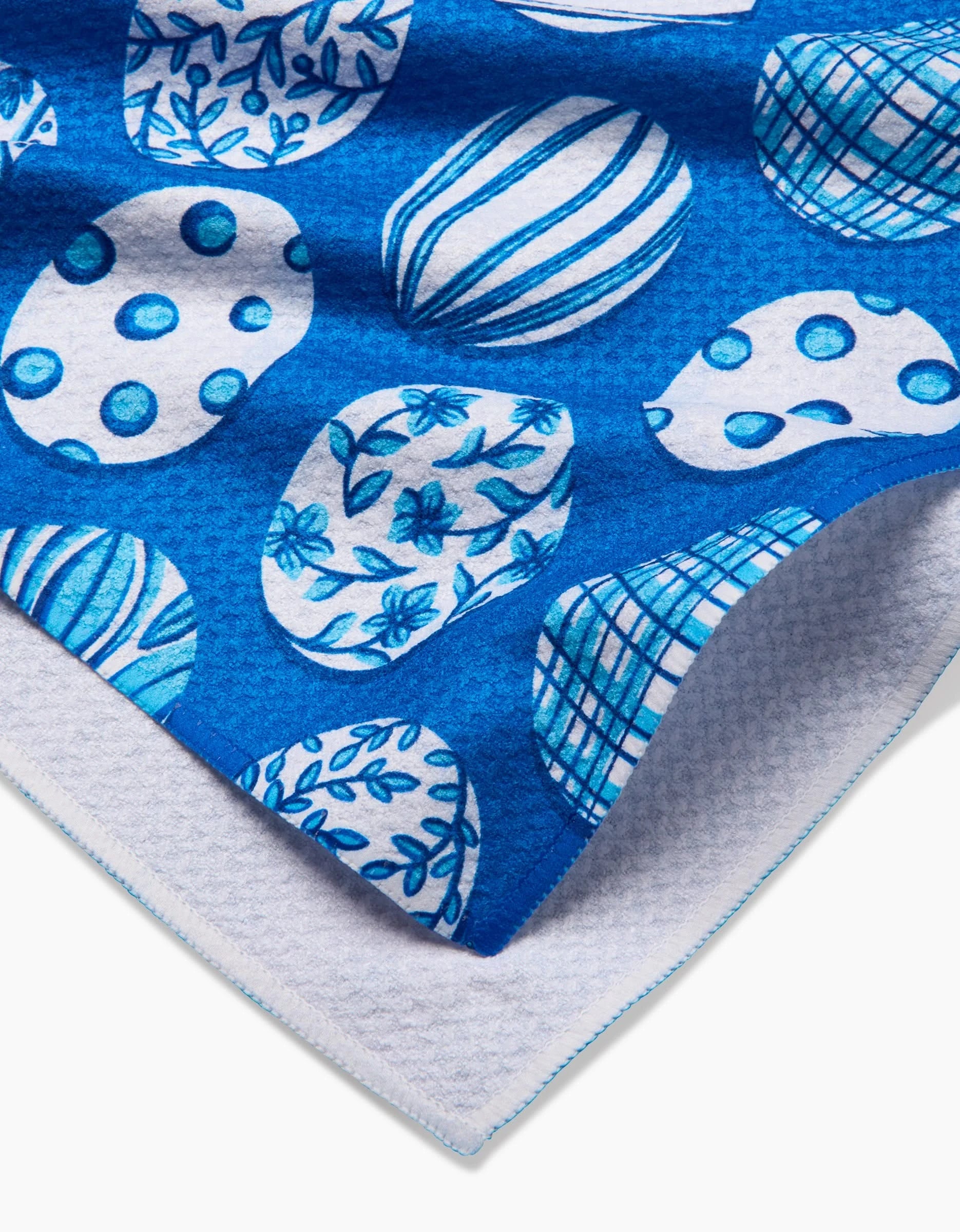 The Delft Blue Easter Eggs tea towel by Sarah Daugherty features blue and white ovals with floral, striped, and polka dot designs. Made from quick-drying recycled materials, it has a textured white underside and blue-stitched edging.