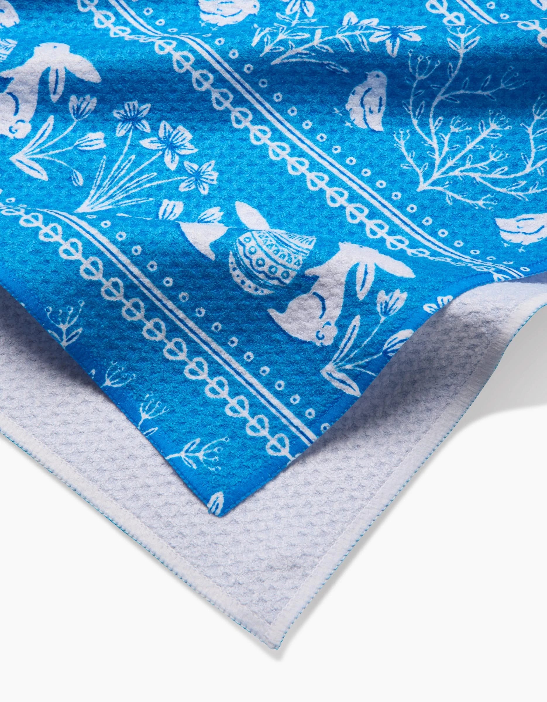A close-up of the Sarah Daugherty Delft Blue Easter Stripe tea towel, featuring white rabbits, flowers, and decorative patterns on blue, layered over light grey textured fabric.