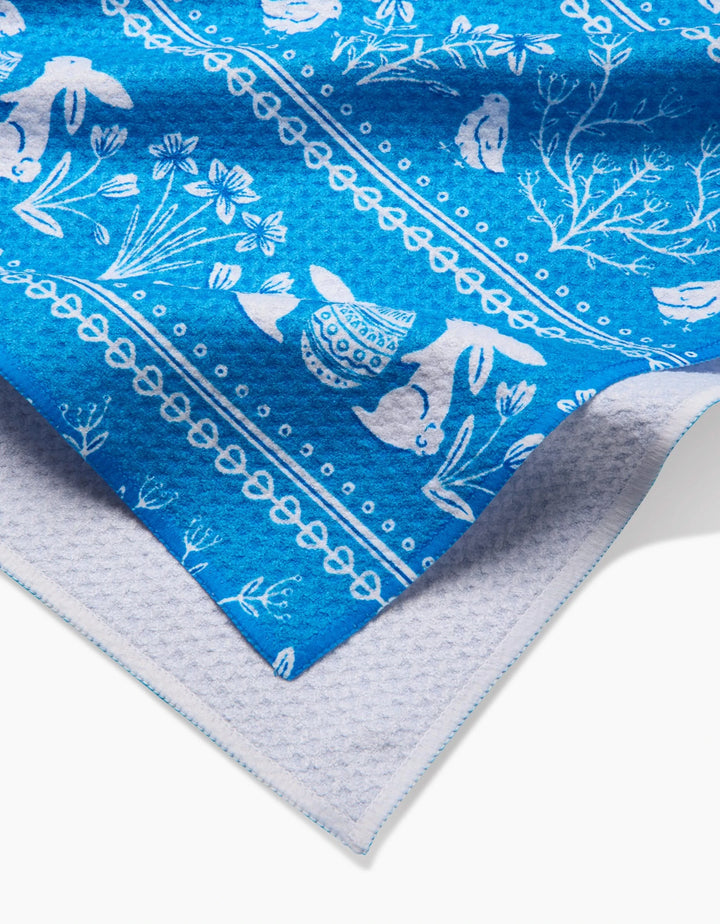 A close-up of the Sarah Daugherty Delft Blue Easter Stripe tea towel, featuring white rabbits, flowers, and decorative patterns on blue, layered over light grey textured fabric.