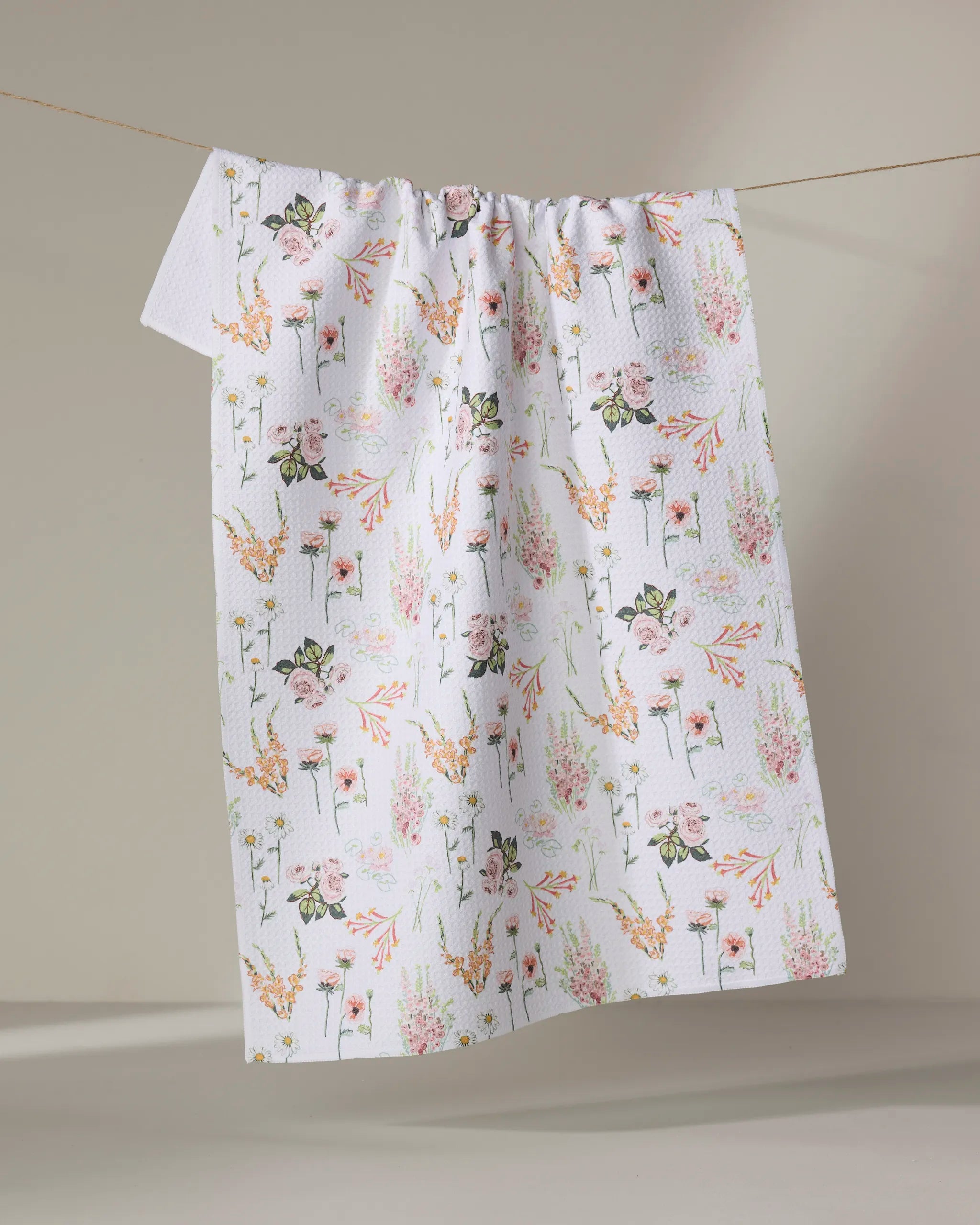 The Elisabeth Wing "Delicate Floral" kitchen towel features pink, orange, and green blossoms on white, hangs on a clothesline against a neutral background. Made from recycled materials—perfect as a tea towel or vibrant accent.