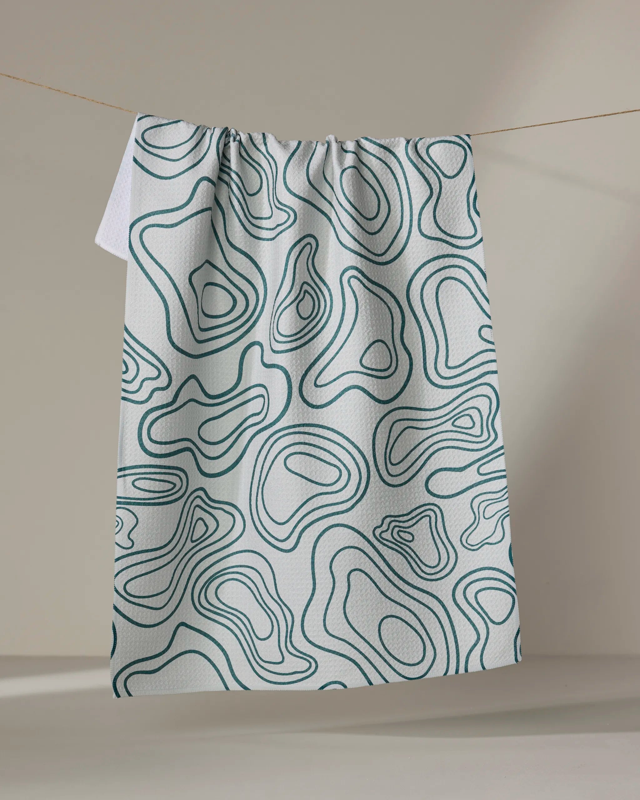 The Depth Chart tea towel by Twig + Spark, made from recycled materials, showcases teal topographic-inspired abstract waves and hangs from a string against a neutral background.