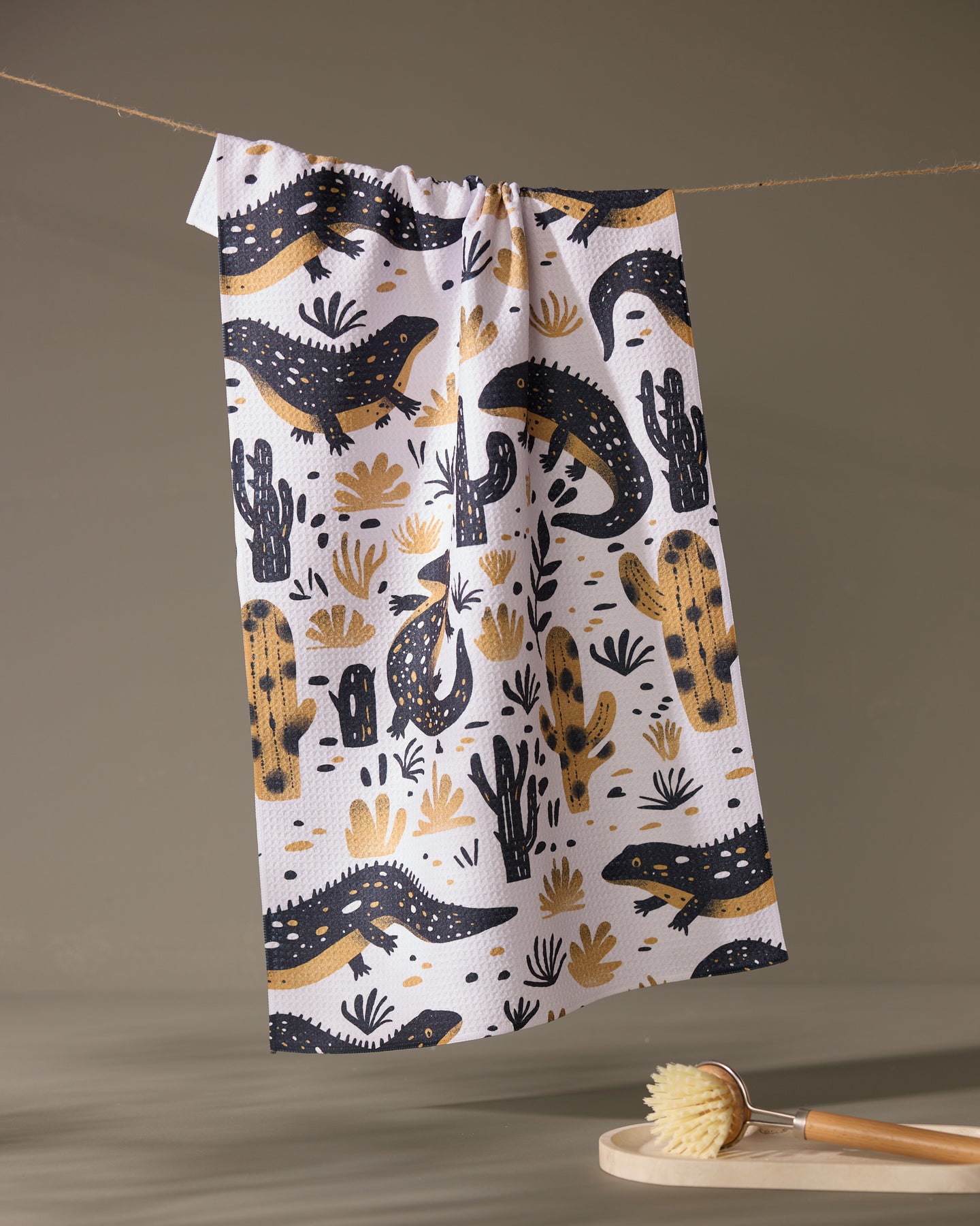The Geometry Desert Dwellers kitchen towel, featuring black and gold lizards and cacti on white, hangs on a clothesline. Below, a wooden dish brush with light bristles rests on a round tray in a softly lit neutral setting.