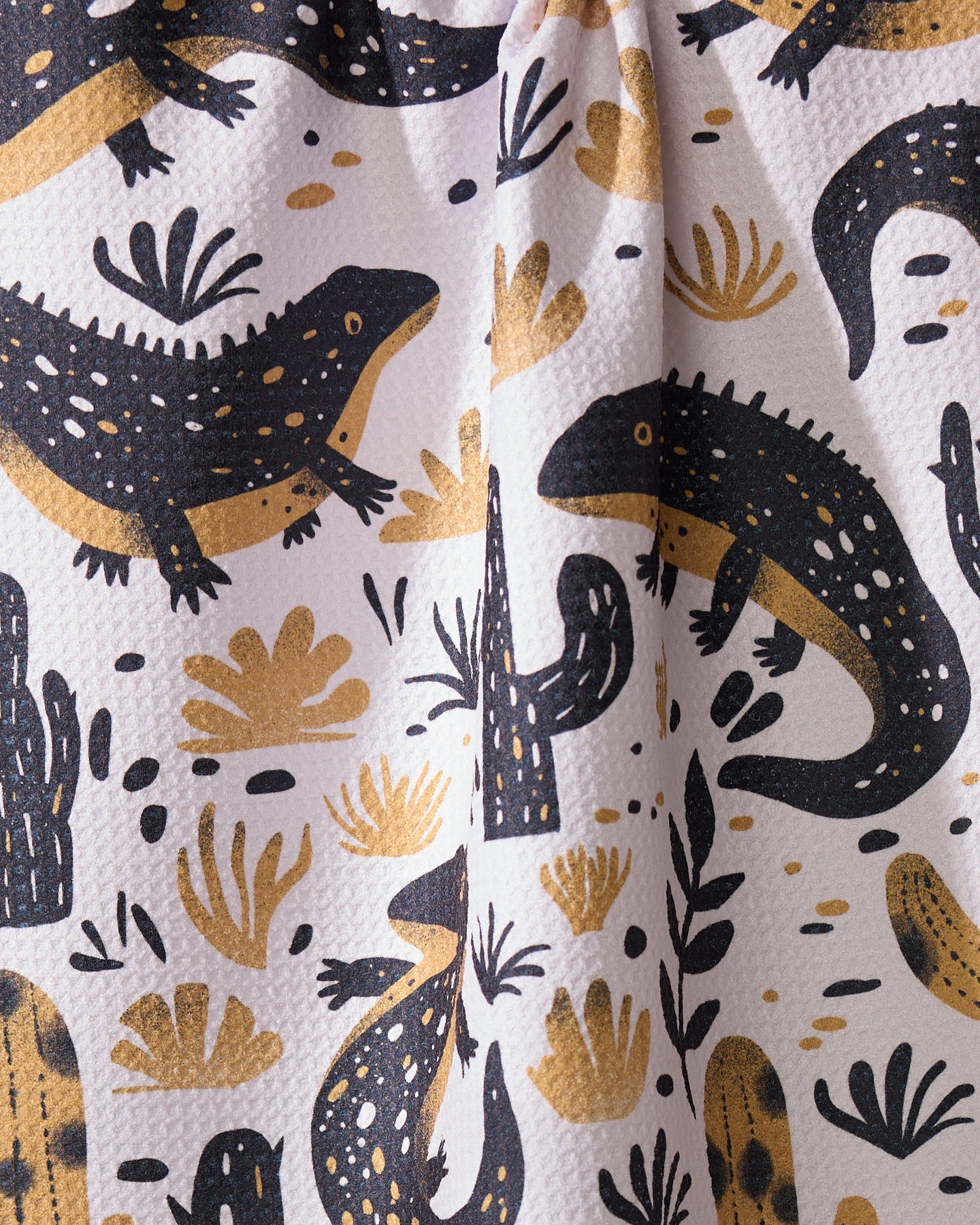 The Desert Dwellers tea towel by Geometry features illustrated black and gold lizards, cacti, and plants on a light, textured background. Made from recycled materials.
